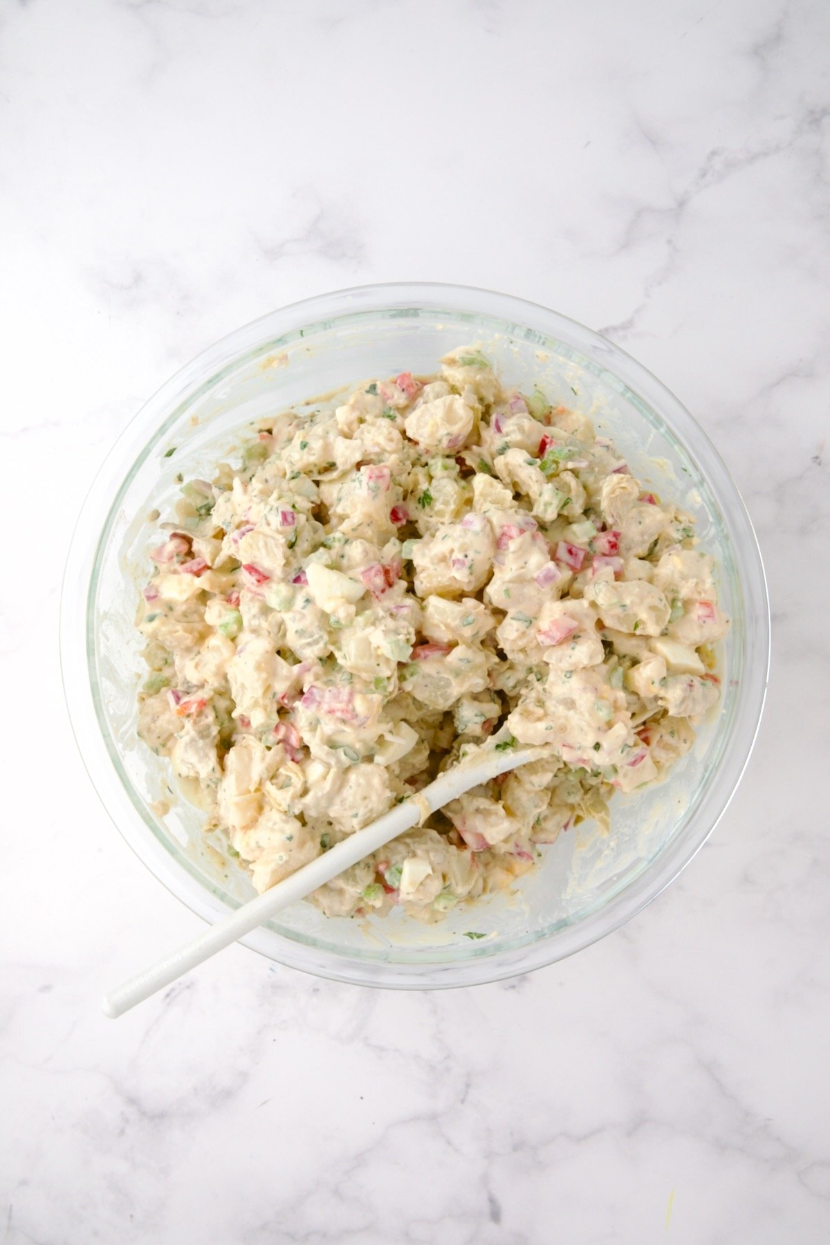 Potato salad in glass bowl with white mixing spoon.
