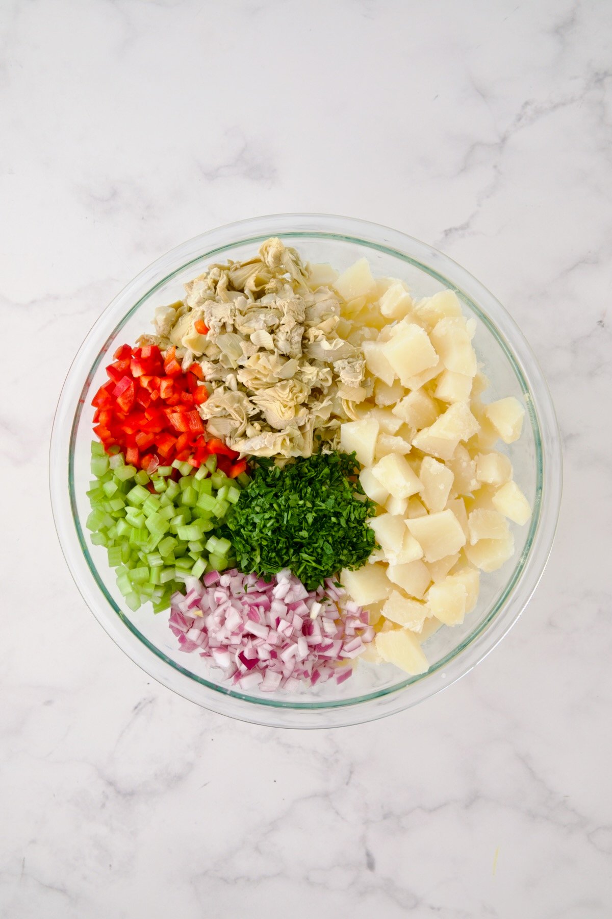 Potatoes, red onion, celery, red bell pepper, artichokes and parsley in glass bowl before being mixed together.