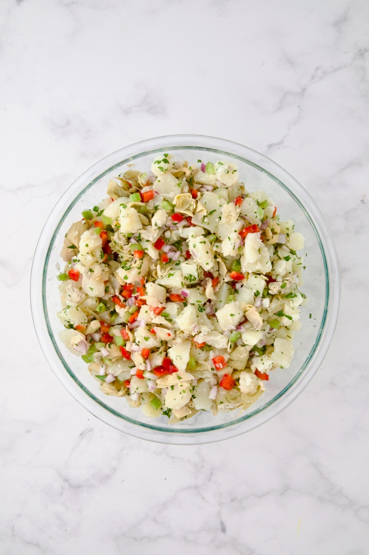 Potatoes, red onion, celery, red bell pepper, artichokes and parsley in glass bowl after being mixed together.