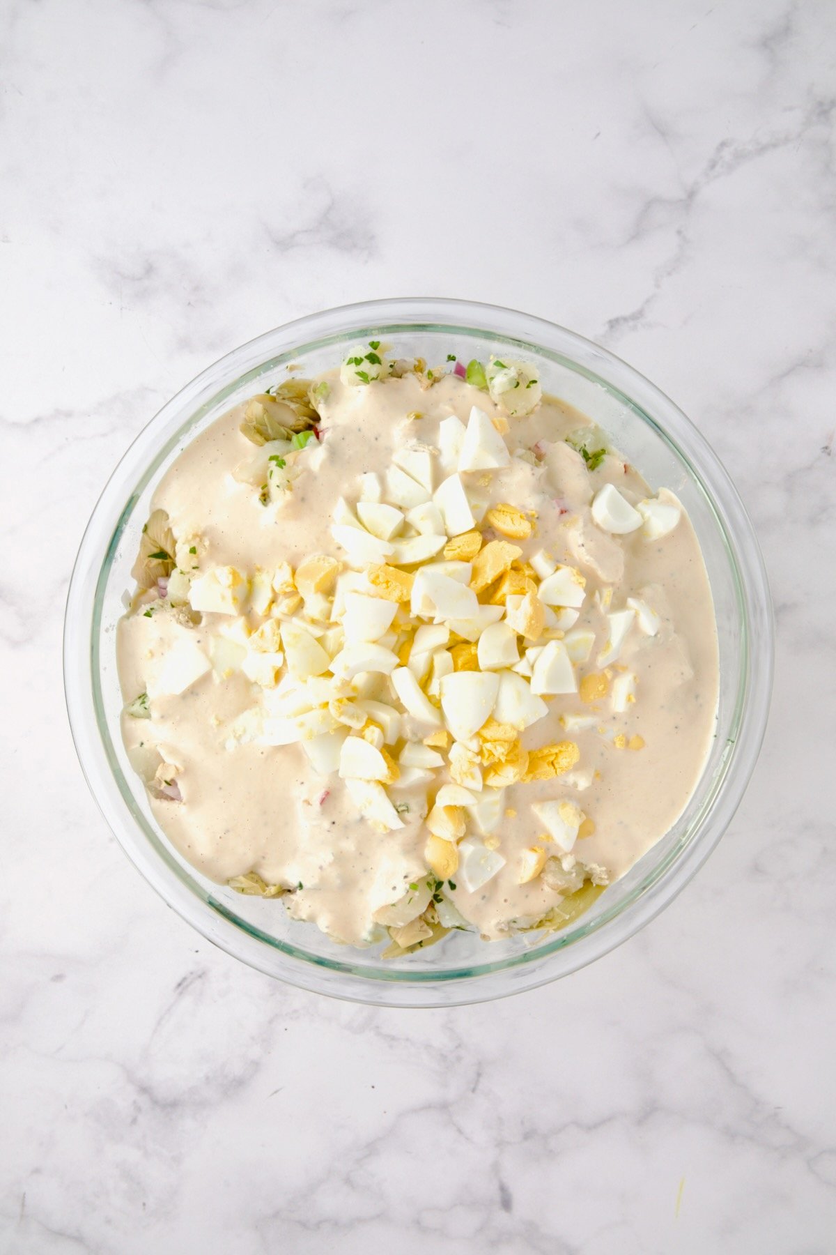 Potatoes, red onion, celery, red bell pepper, artichokes, and parsley in a glass bowl after being mixed together with dressing and eggs added.