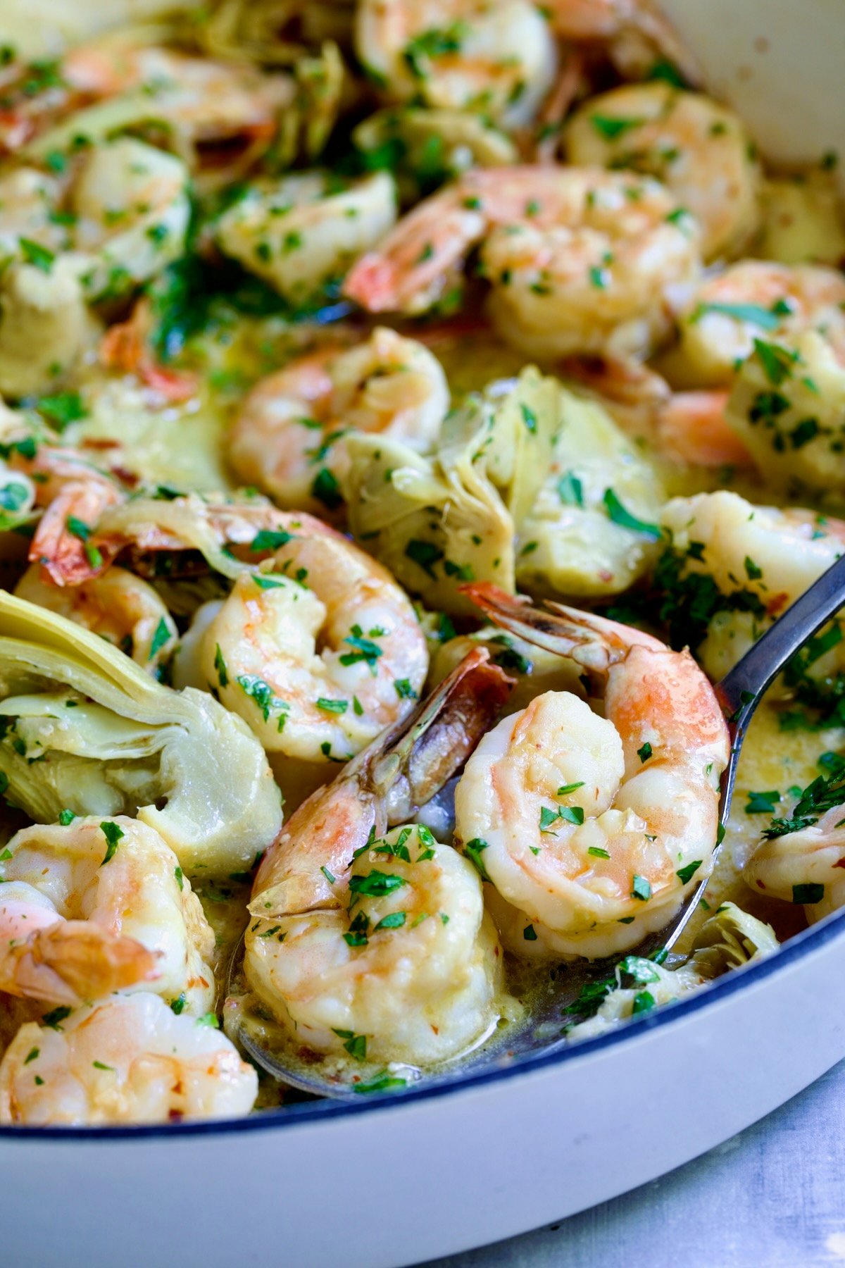 Shrimp Scampi with Artichokes in white enamel cast iron pan with serving spoon scooping two shrimp up.