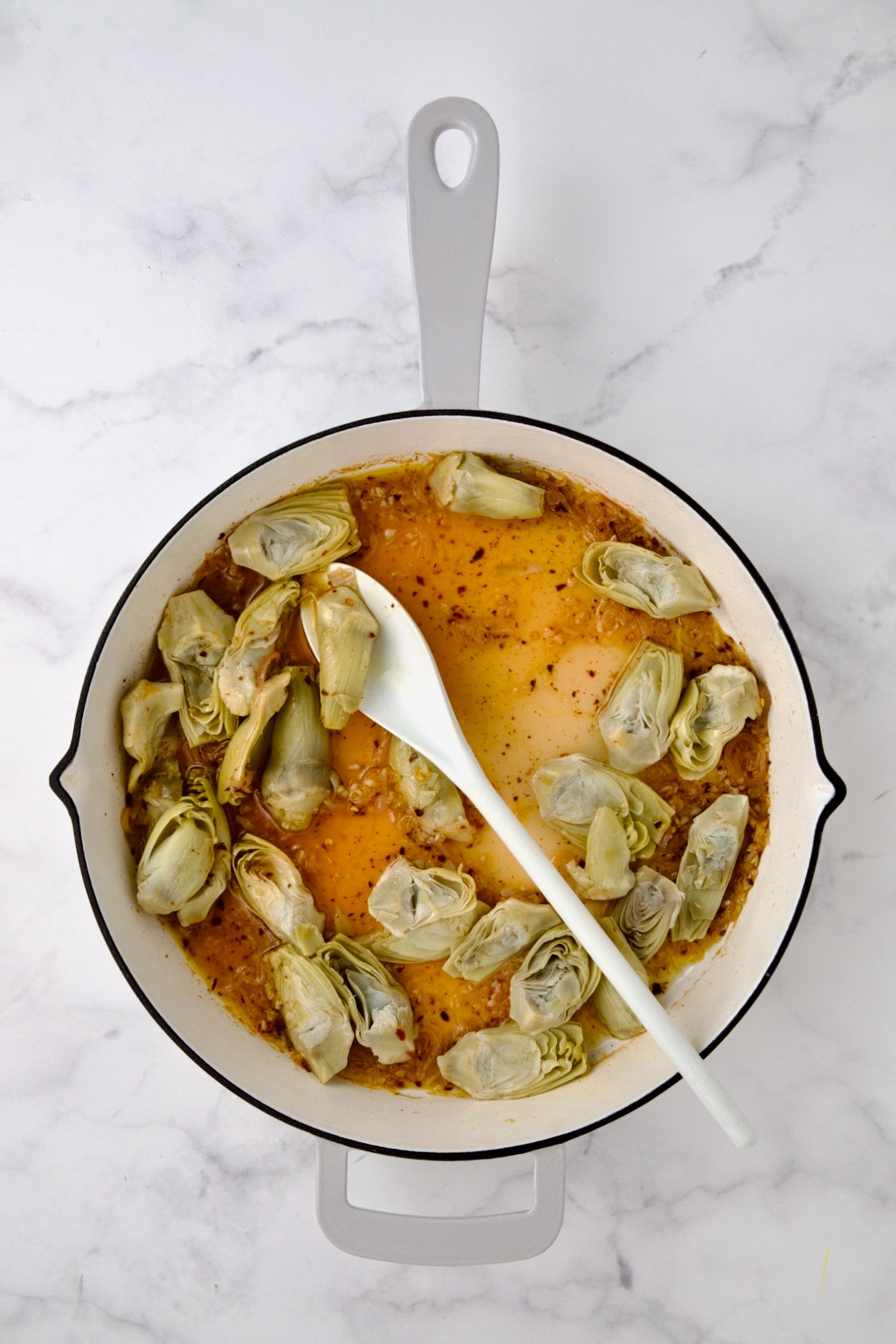 Artichokes added to wine, lemon and shallots in white enamel cast iron pan.