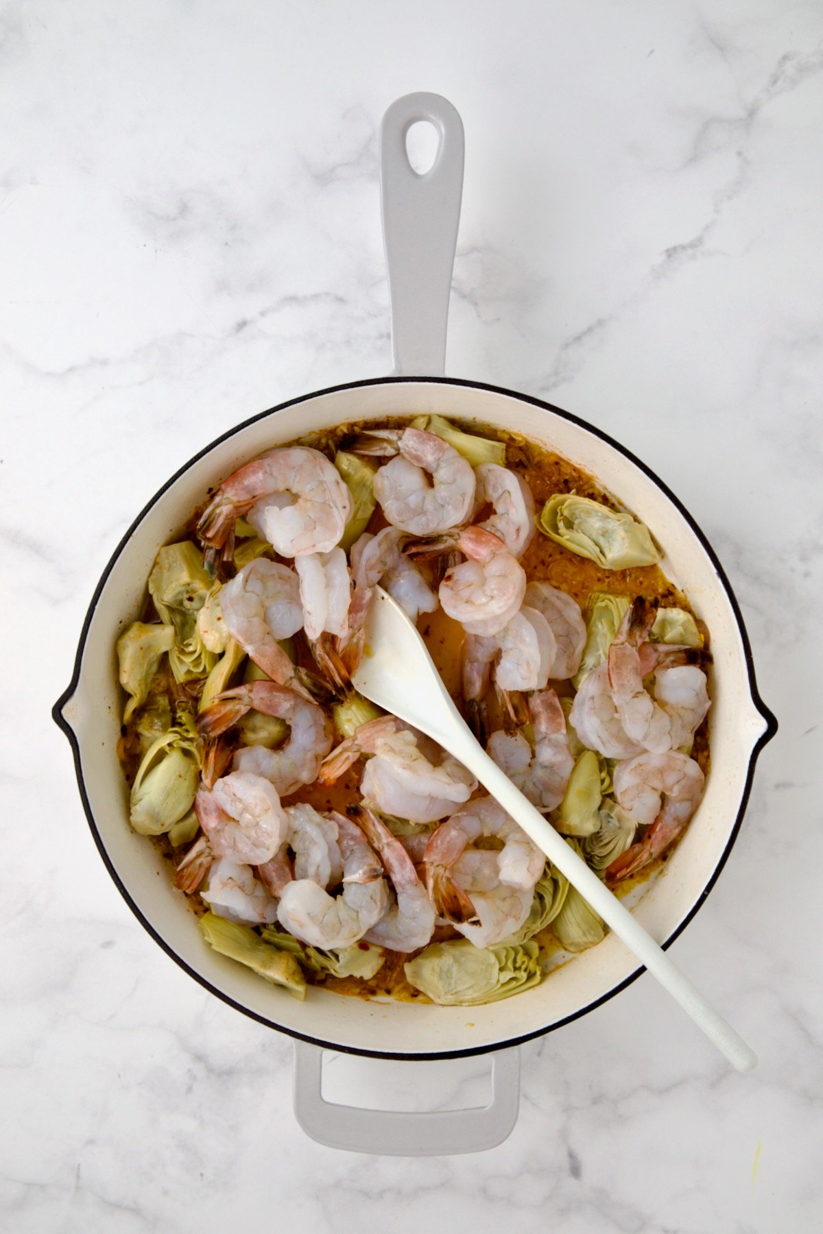 Shrimp added to artichokes in white enamel cast iron pan being stirred with white mixing spoon.