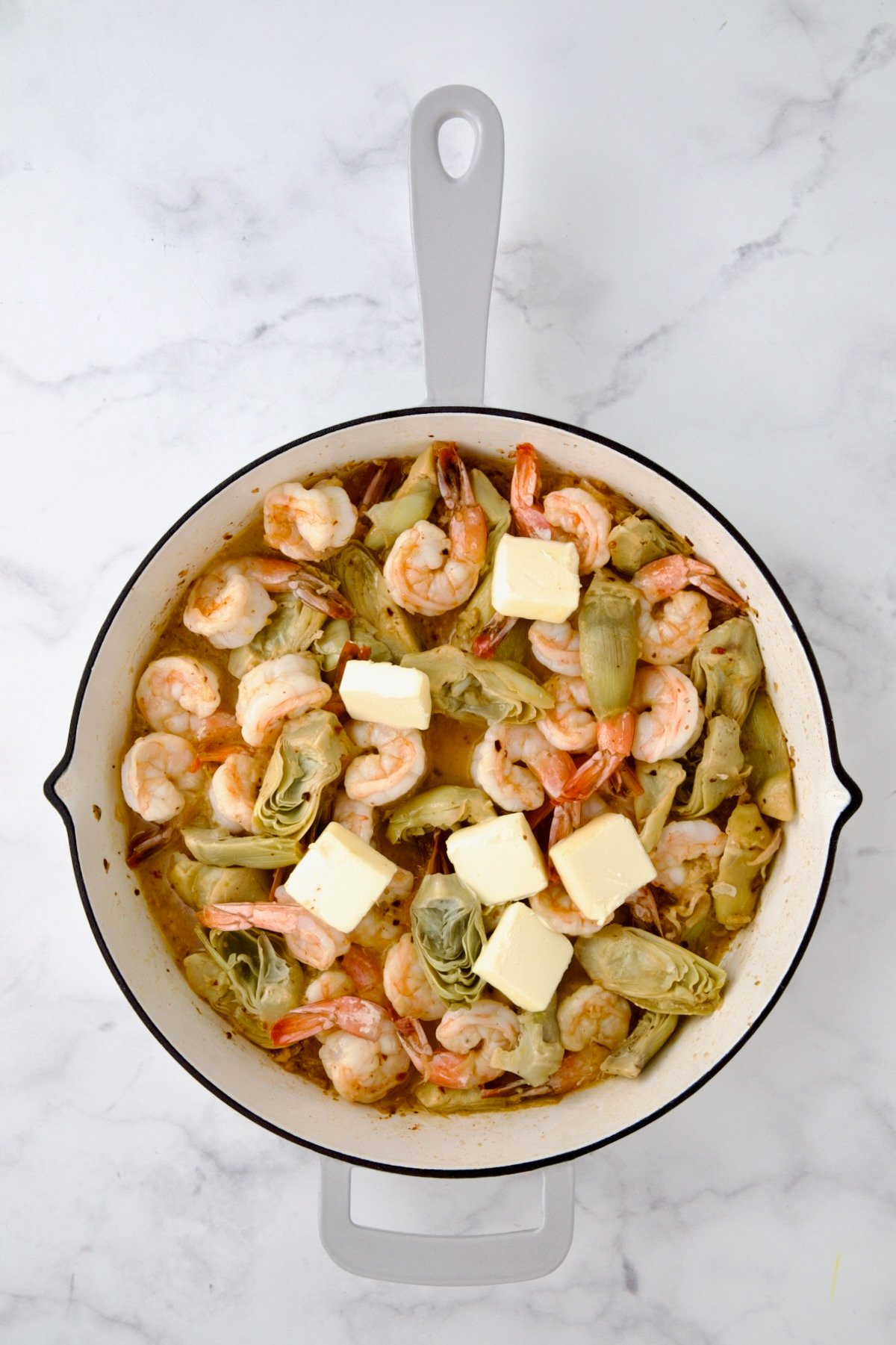 Butter added cooked shrimp and artichokes in white enamel cast iron pan.