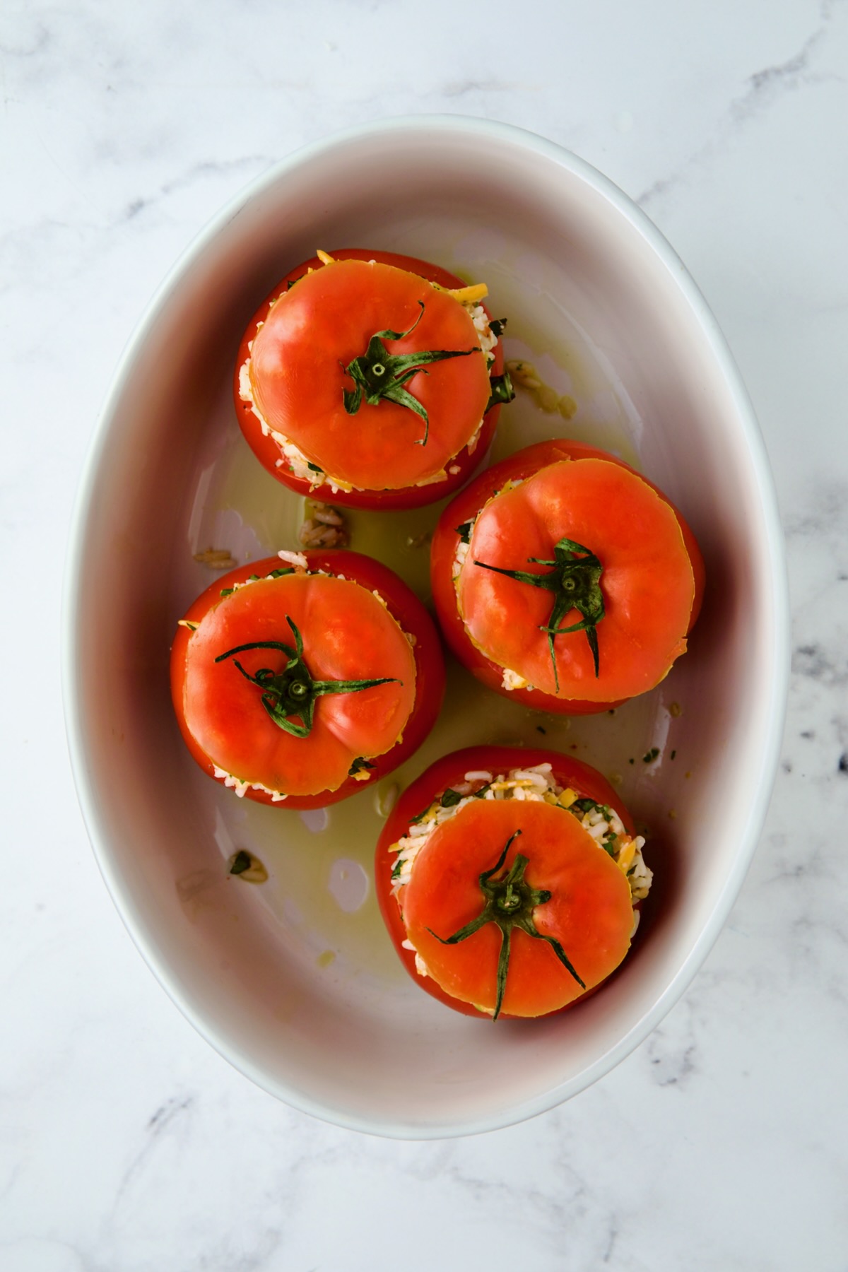 Four round cheese and rice-stuffed tomatoes in oval baking dish before being baked with the tomato tops placed back on top.