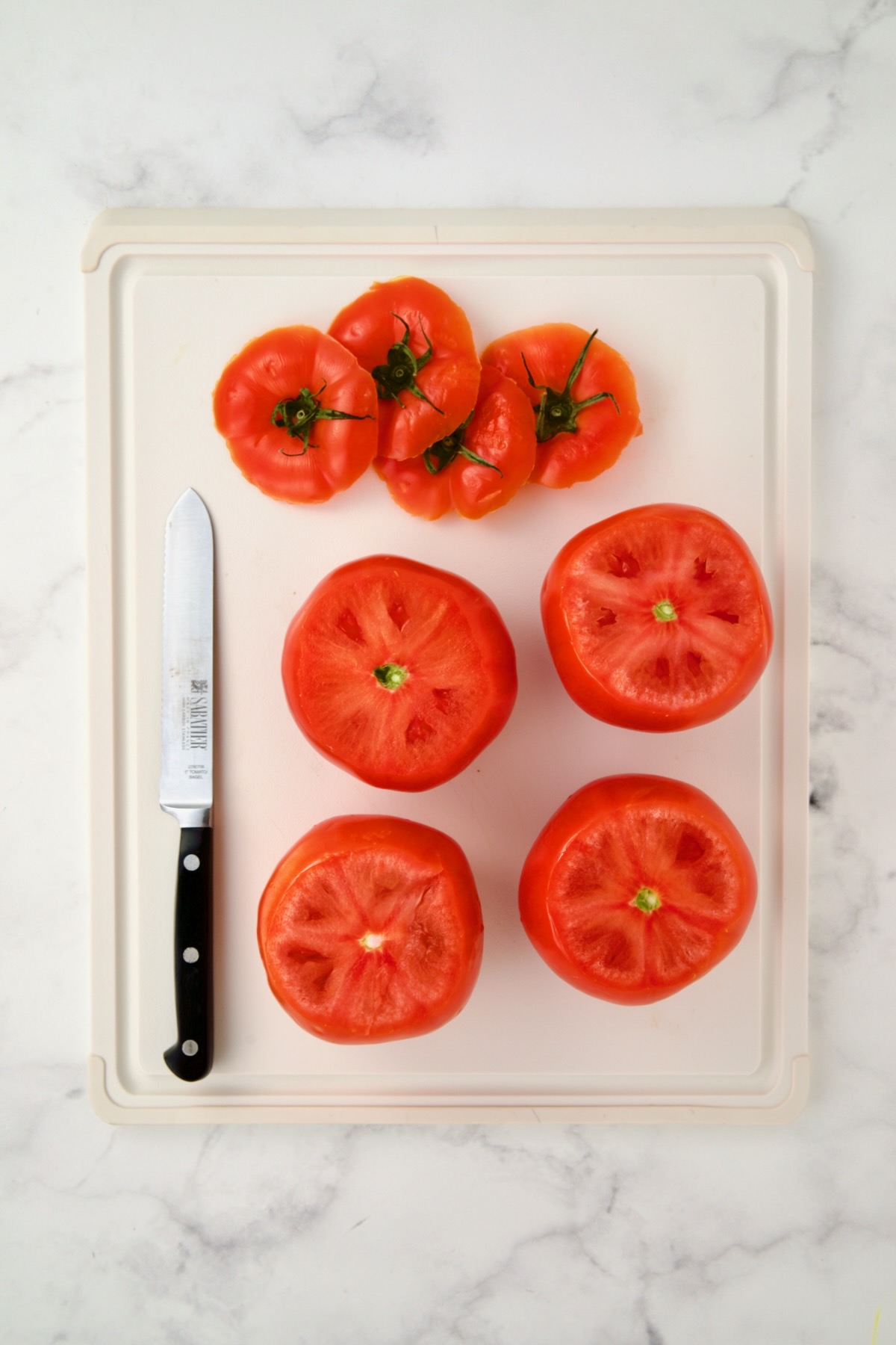 Four round tomatoes with the tops sliced off on white plastic cutting board.
