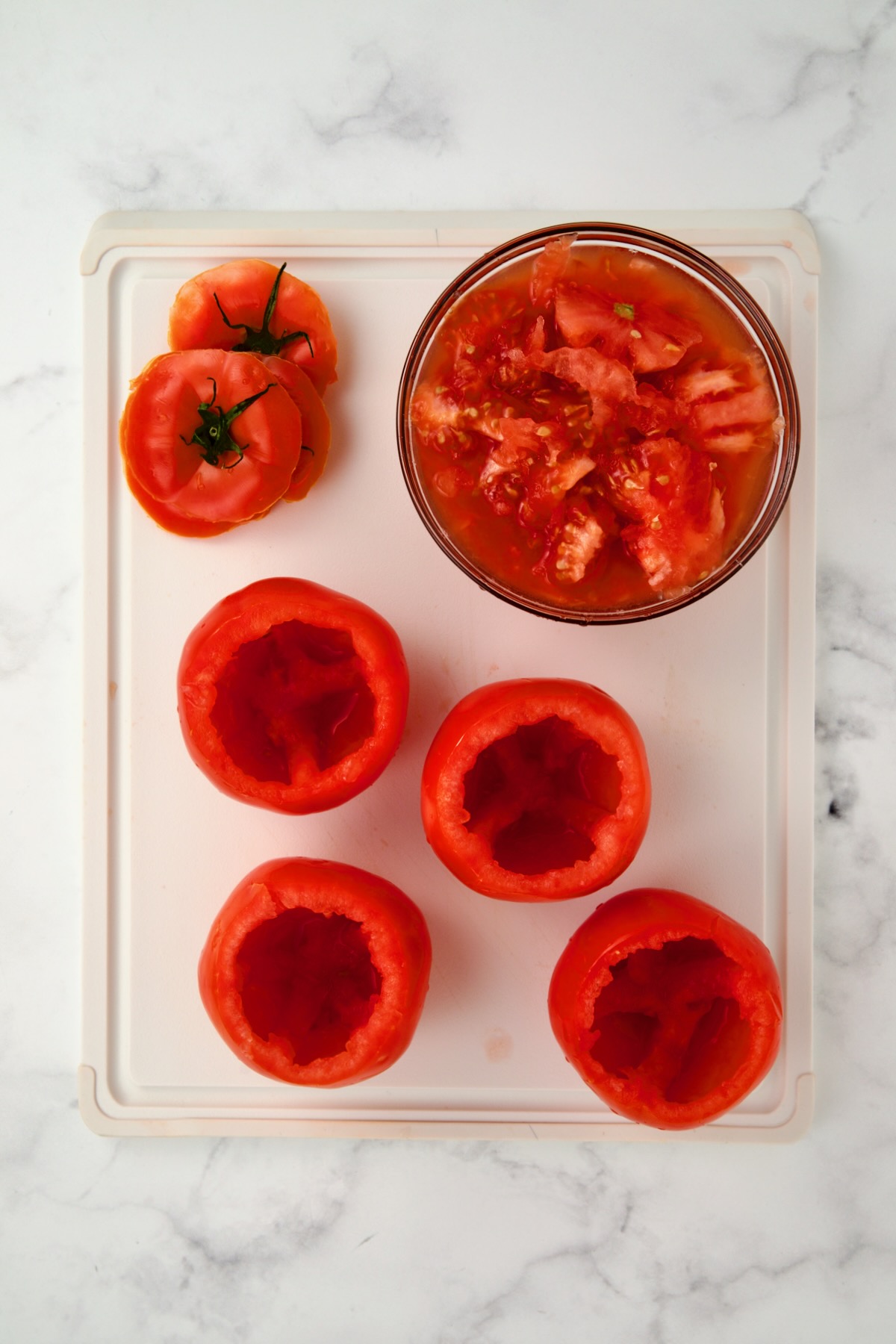 Four hollowed out round tomatoes with the lids and the pulp in a glass bowl on a white plastic cutting board.