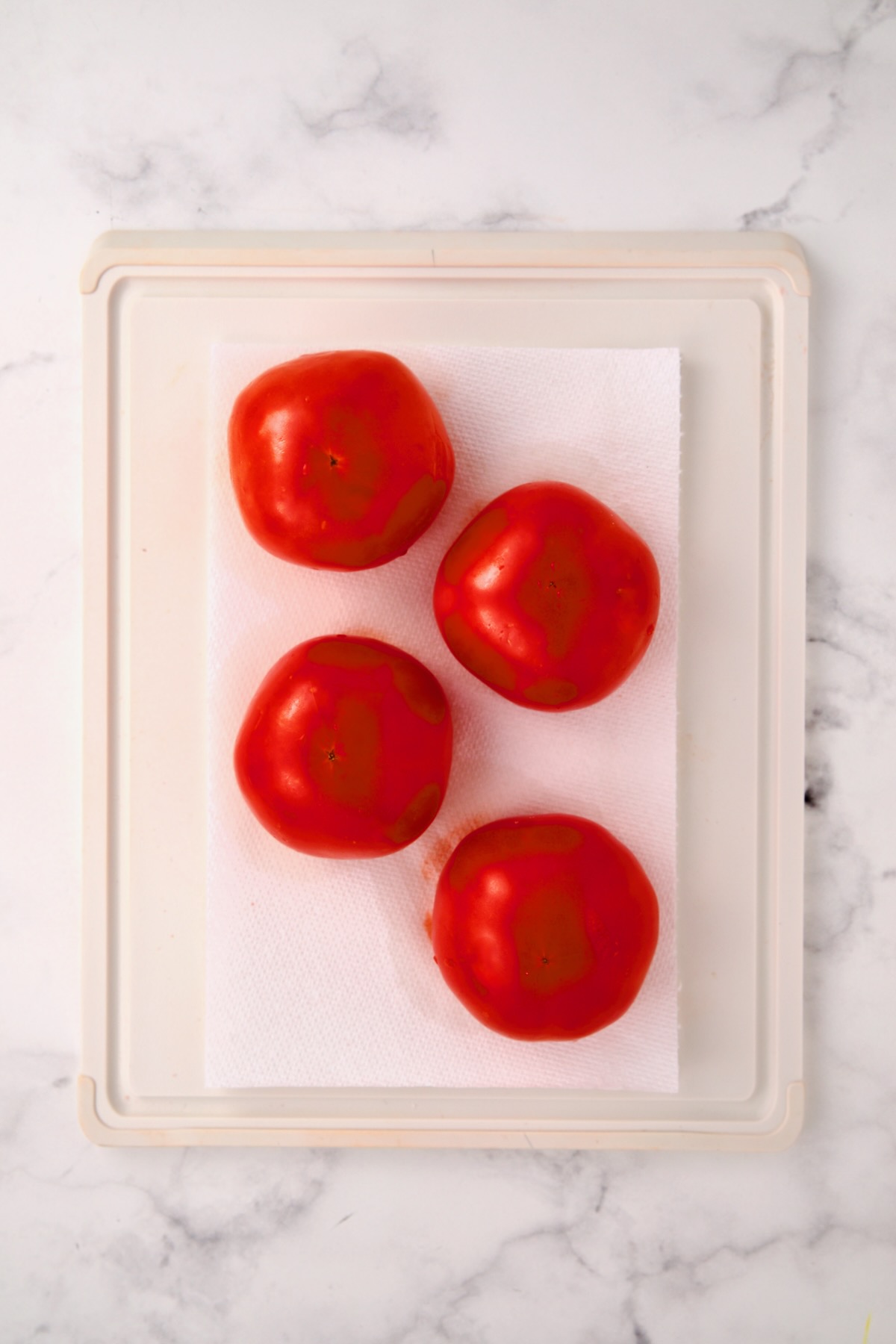 Four round tomatoes inverted onto paper towels on white plastic cutting board.