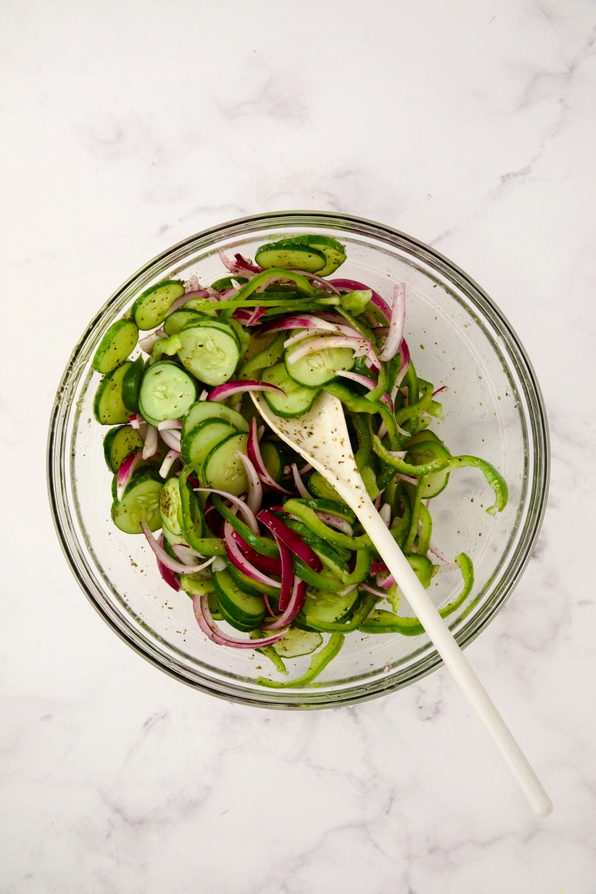 Sliced onion, green pepper and cucumbers in glass bowl being stirred with white mixing spoon.