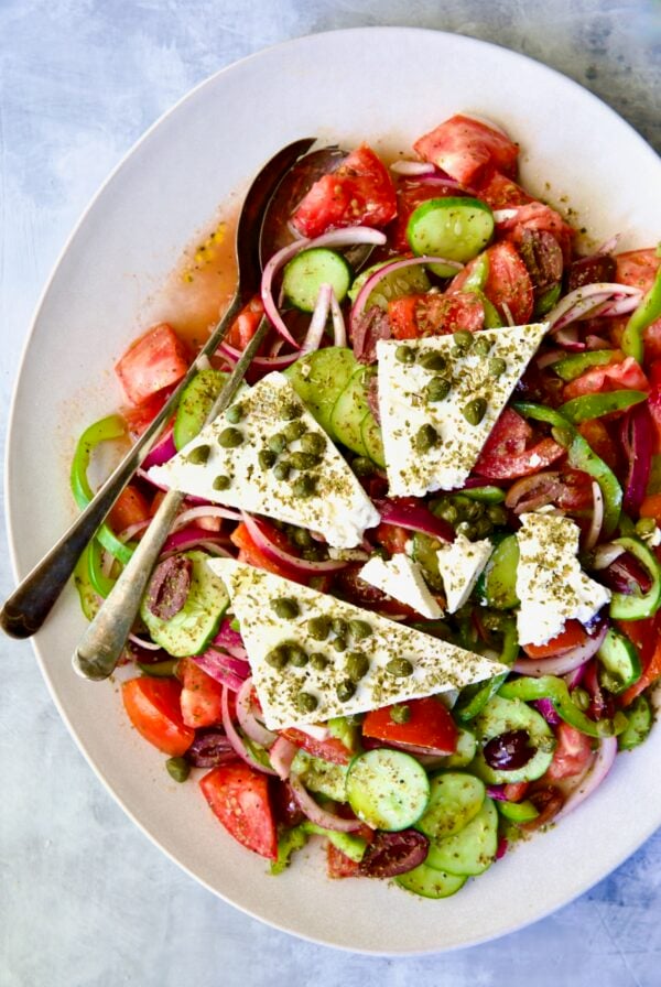 Classic Greek Salad with broken chunks of feta triangles on white oval platter with serving utensils.