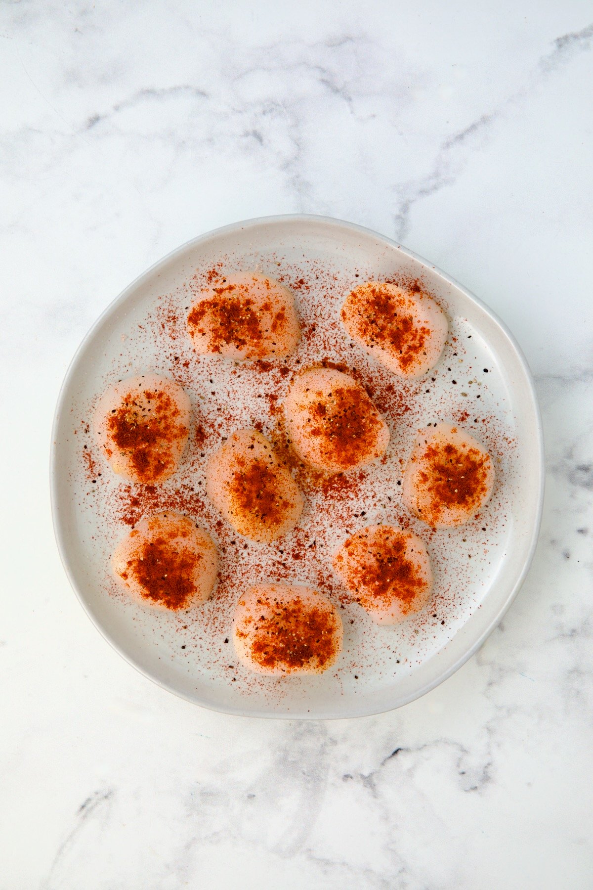 Uncooked seasoned sea scallops on white plate.