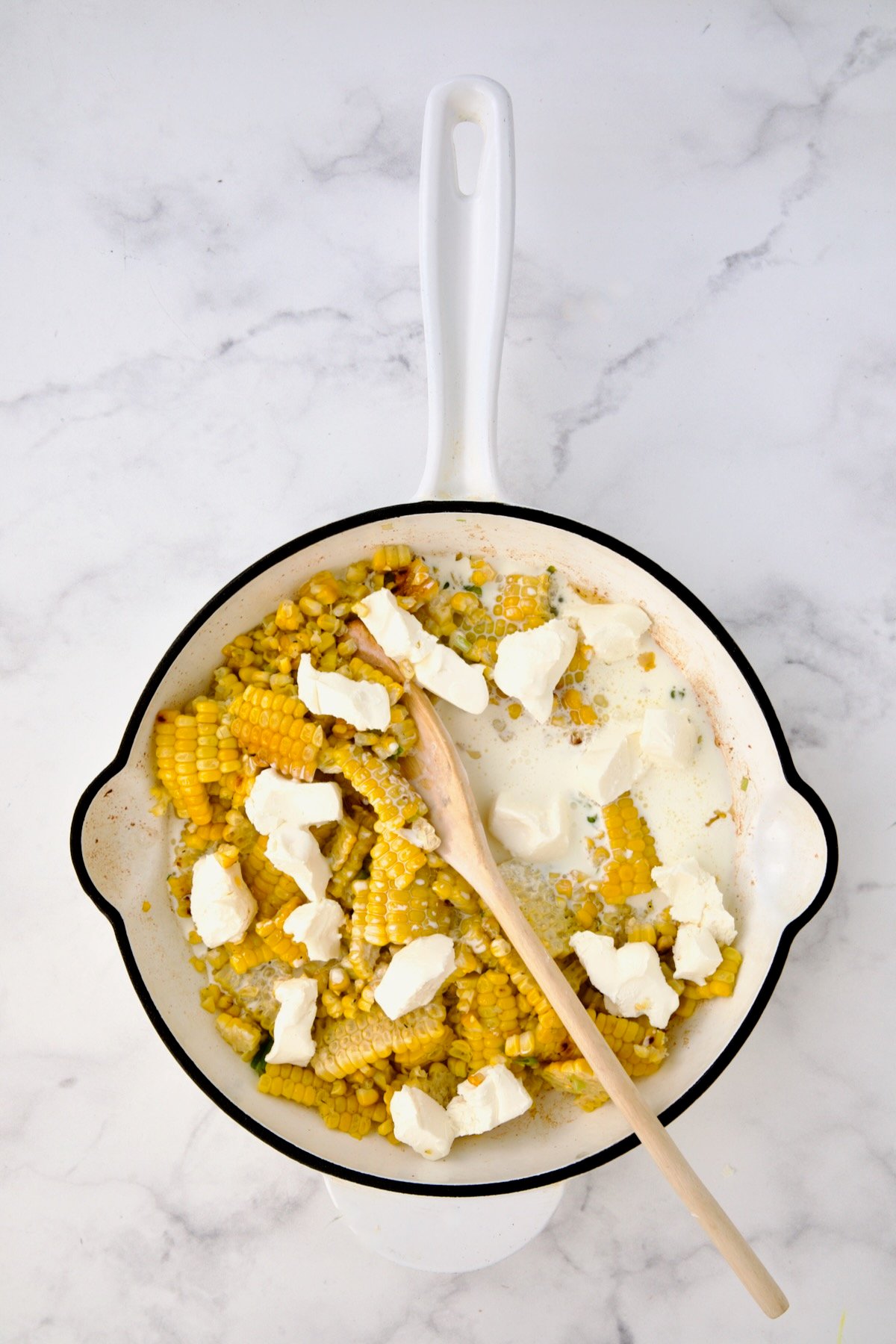 Cream cheese and heavy cream added to corn in white enamel cast iron skillet before being stirred in.