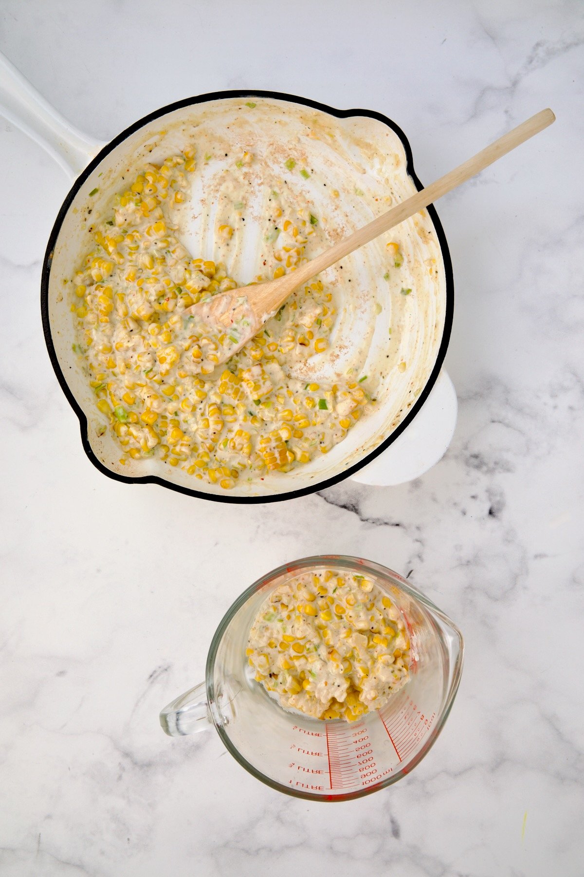 Creamy corn combination in white enamel cast iroon skillet with some placed in a glass measuring cup.