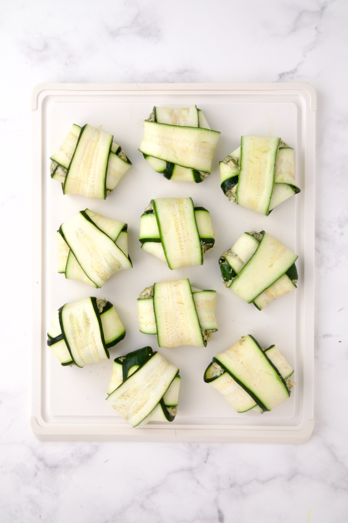 Ten fully assembled zucchini ravioli on white cutting board.
