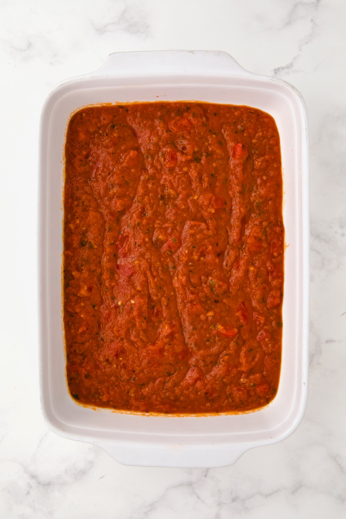 Marinara sauce in the bottom of a rectangular white ceramic baking dish.