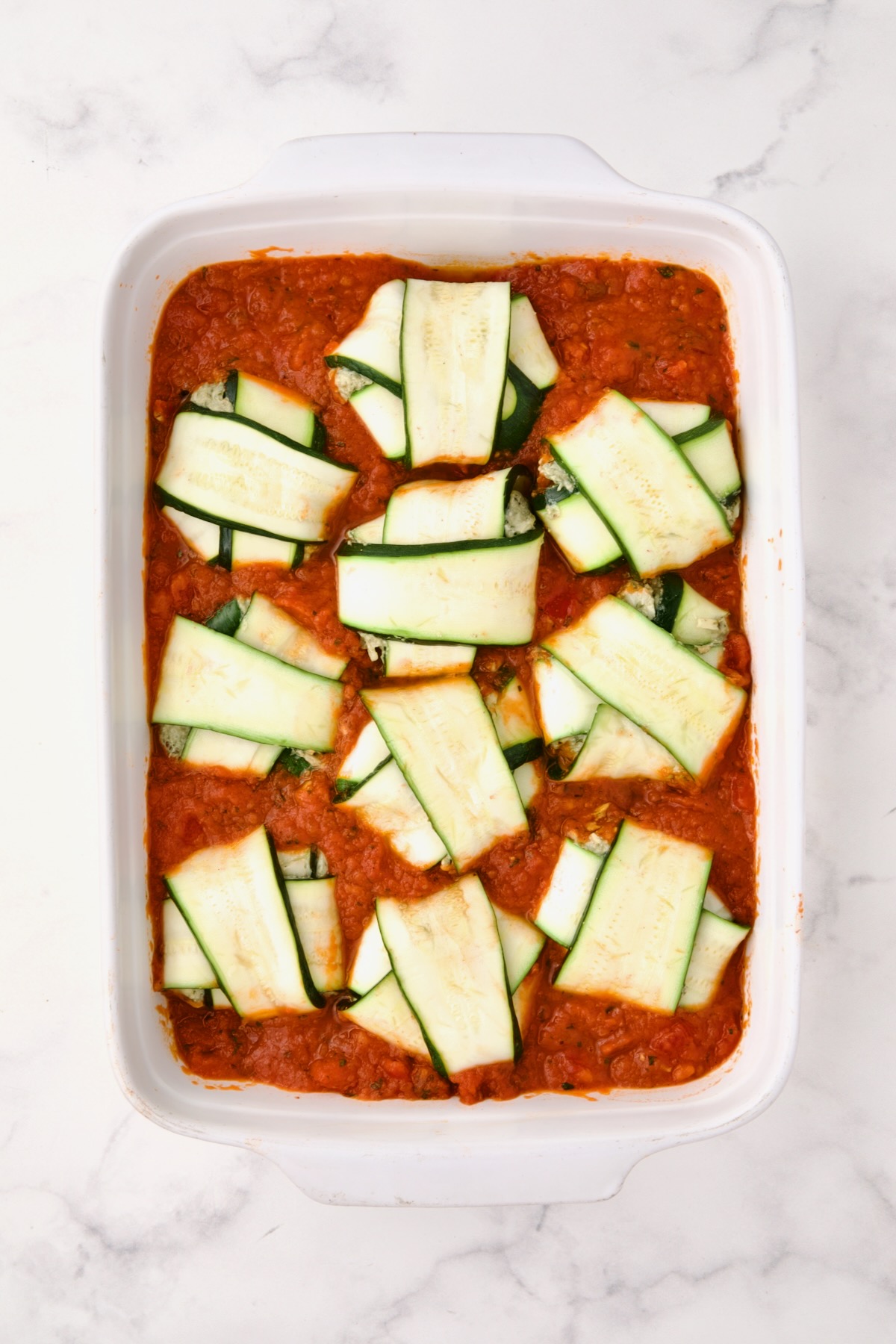 Ten assembled ravioli placed on the marinara sauce in a rectangular white ceramic baking dish before cheese is placed on the top.