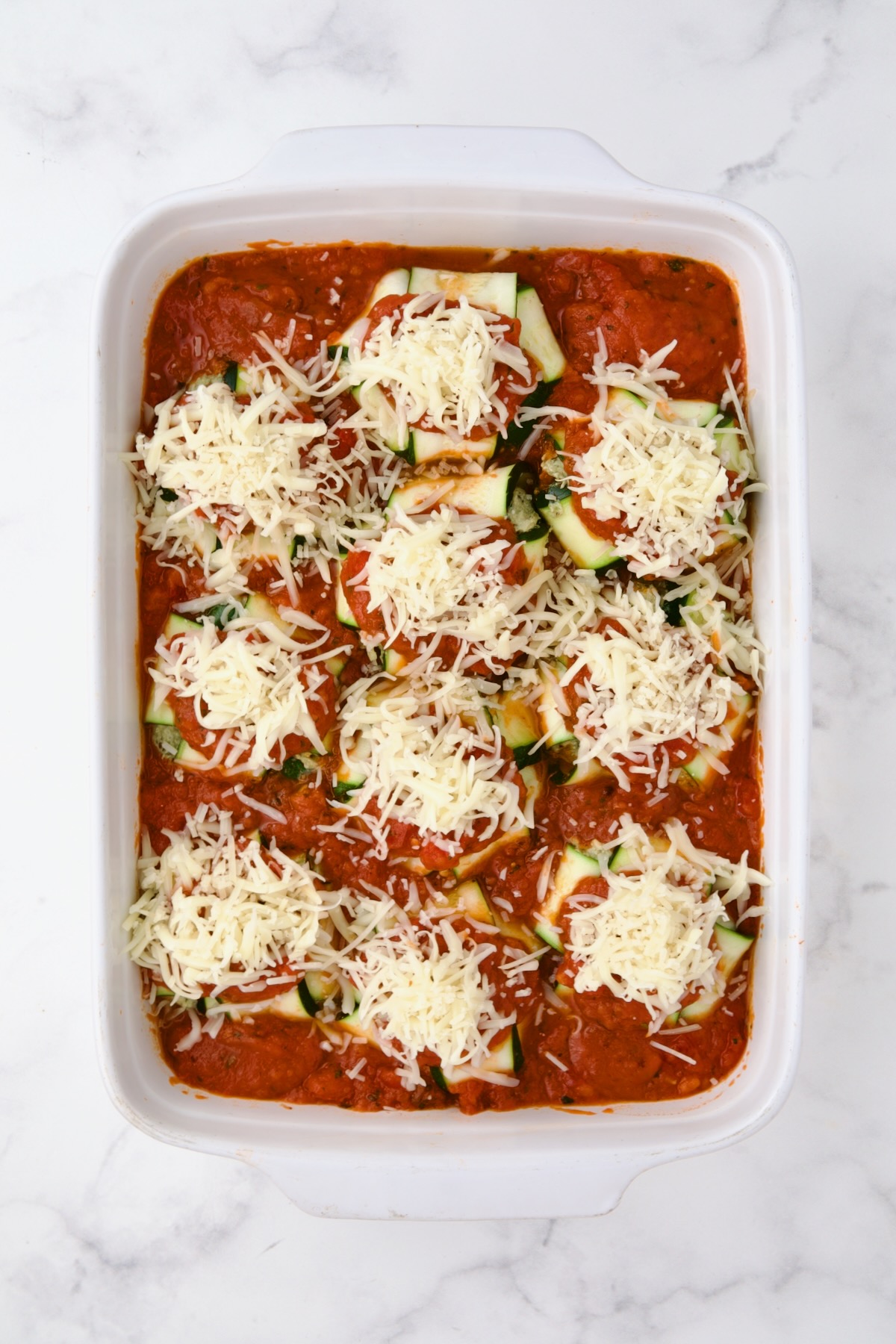 Fully assembled zucchini ravioli with cheese added before being baked in rectangular white ceramic baking dish.