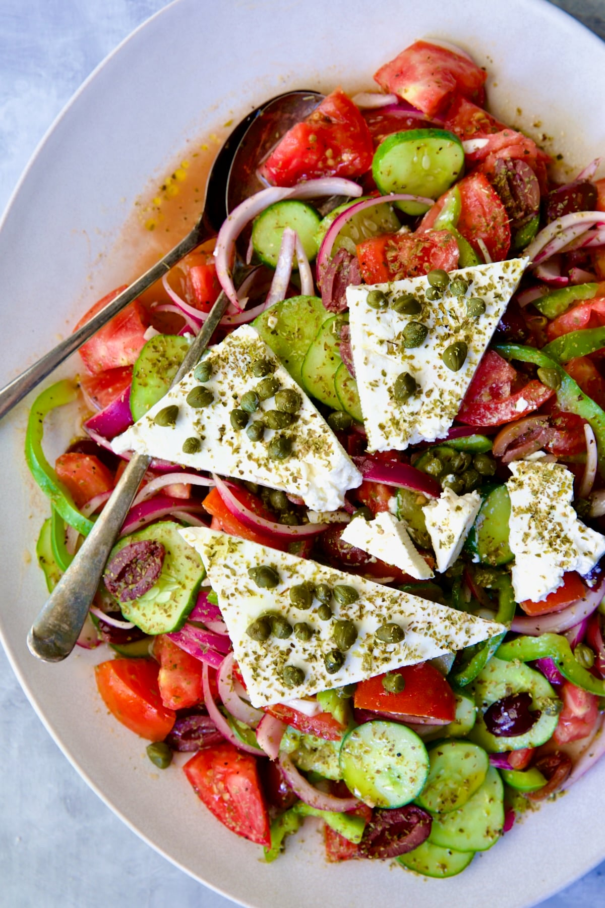 Classic Greek Salad with broken chunks of feta triangles on white oval platter with serving utensils.
