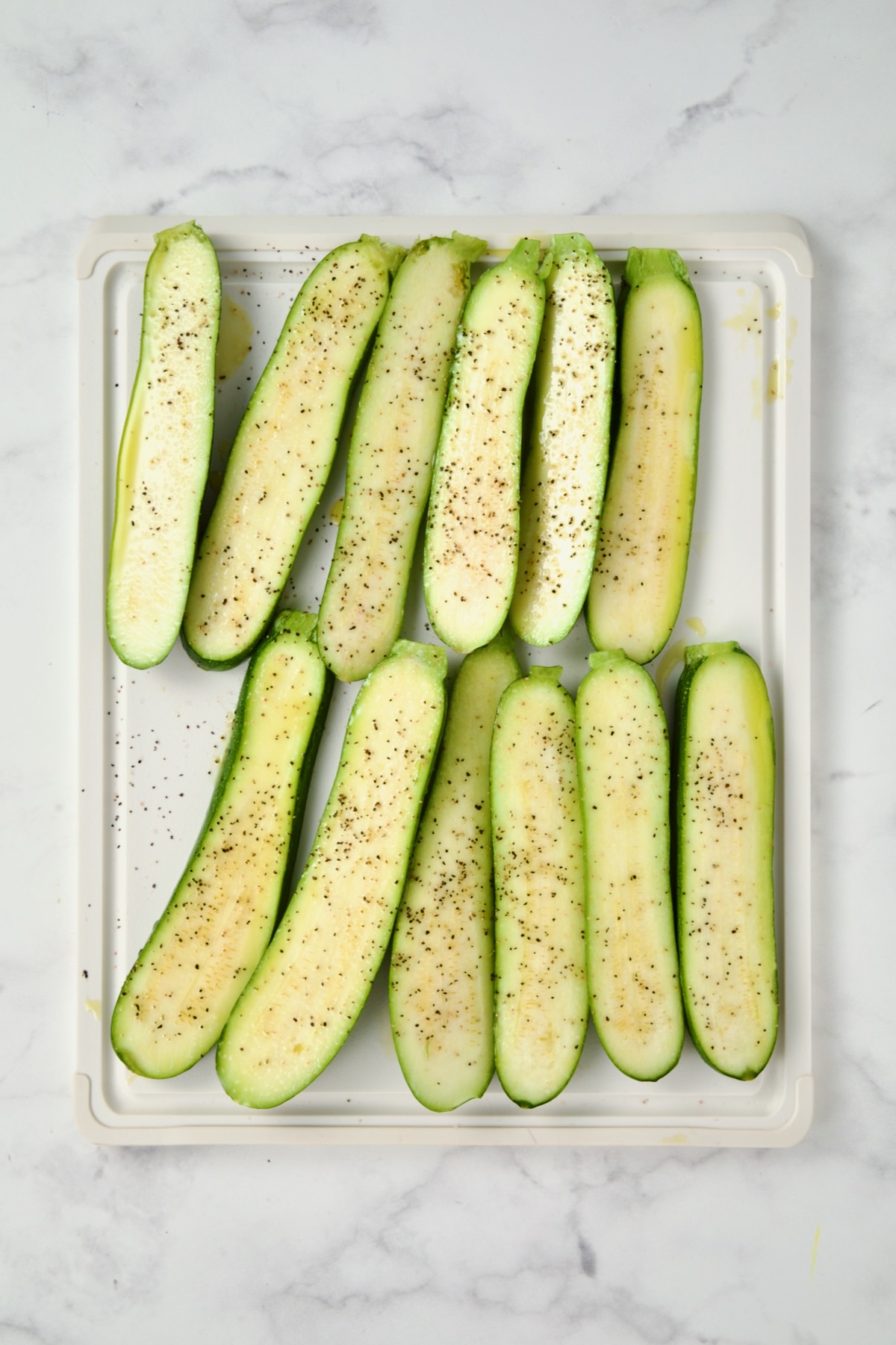 Sliced zucchini on white plastic cutting board seasoned with salt, pepper and drizzled with olive oil.