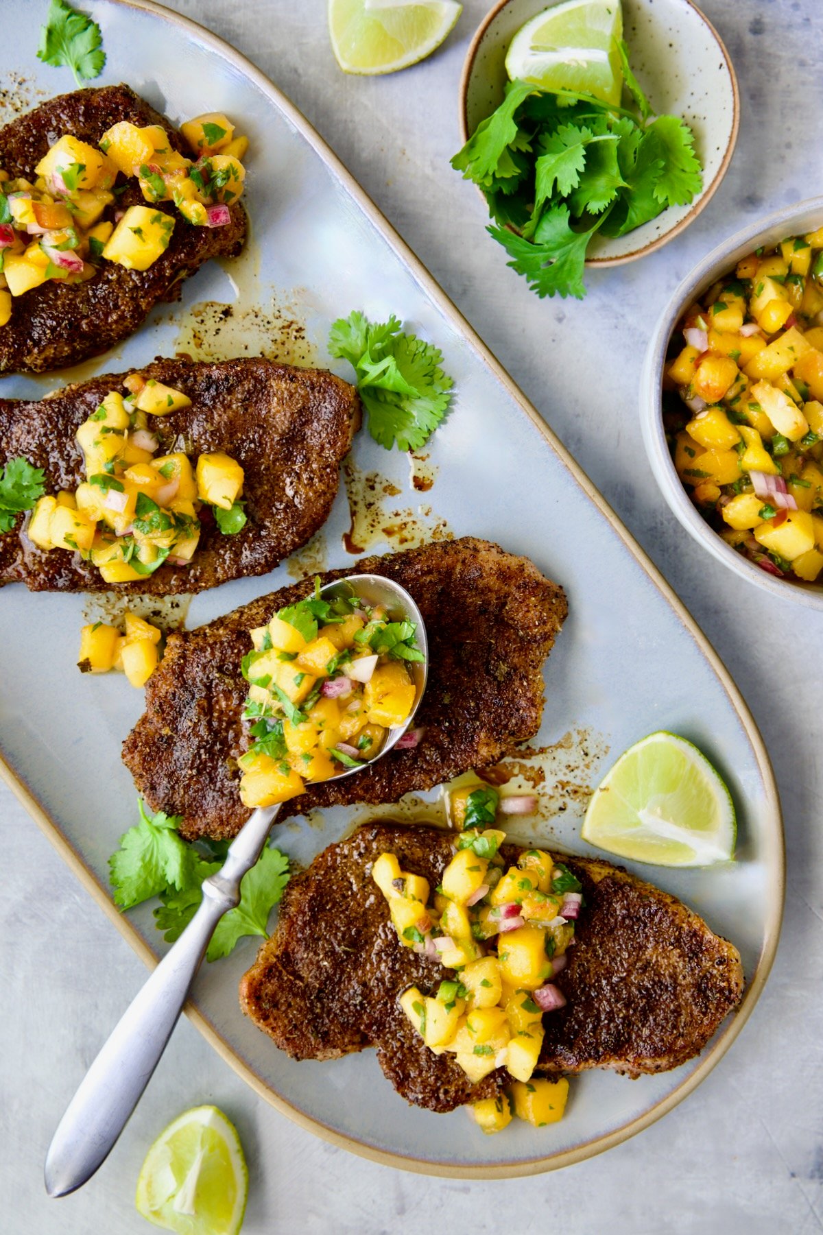 Four grilled Pork Chops with Peach Salsa on oval gray platter with salsa-filled spoon resting on one of the chops.