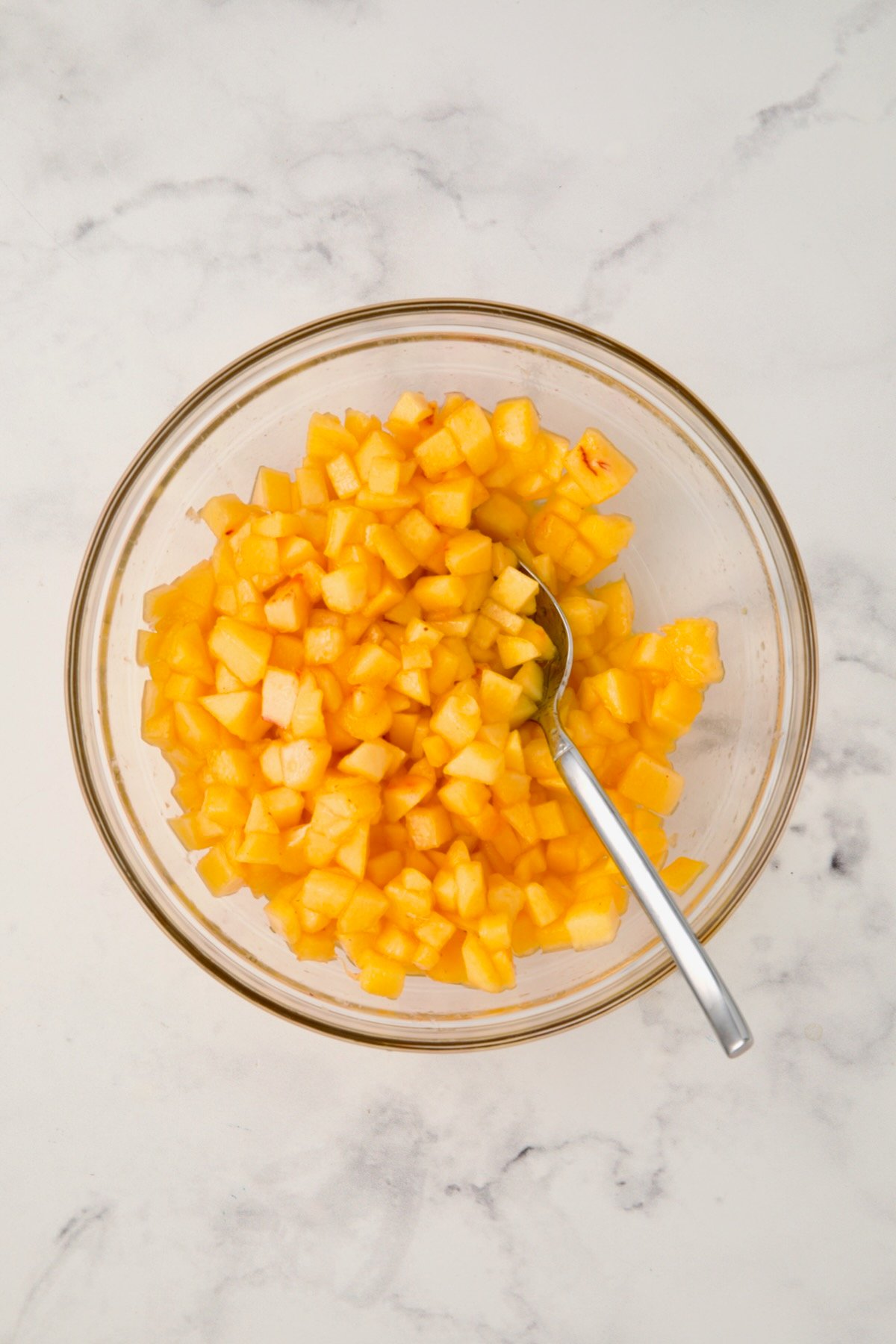 Diced peaches in glass bowl being stirred with stainless steel spoon.