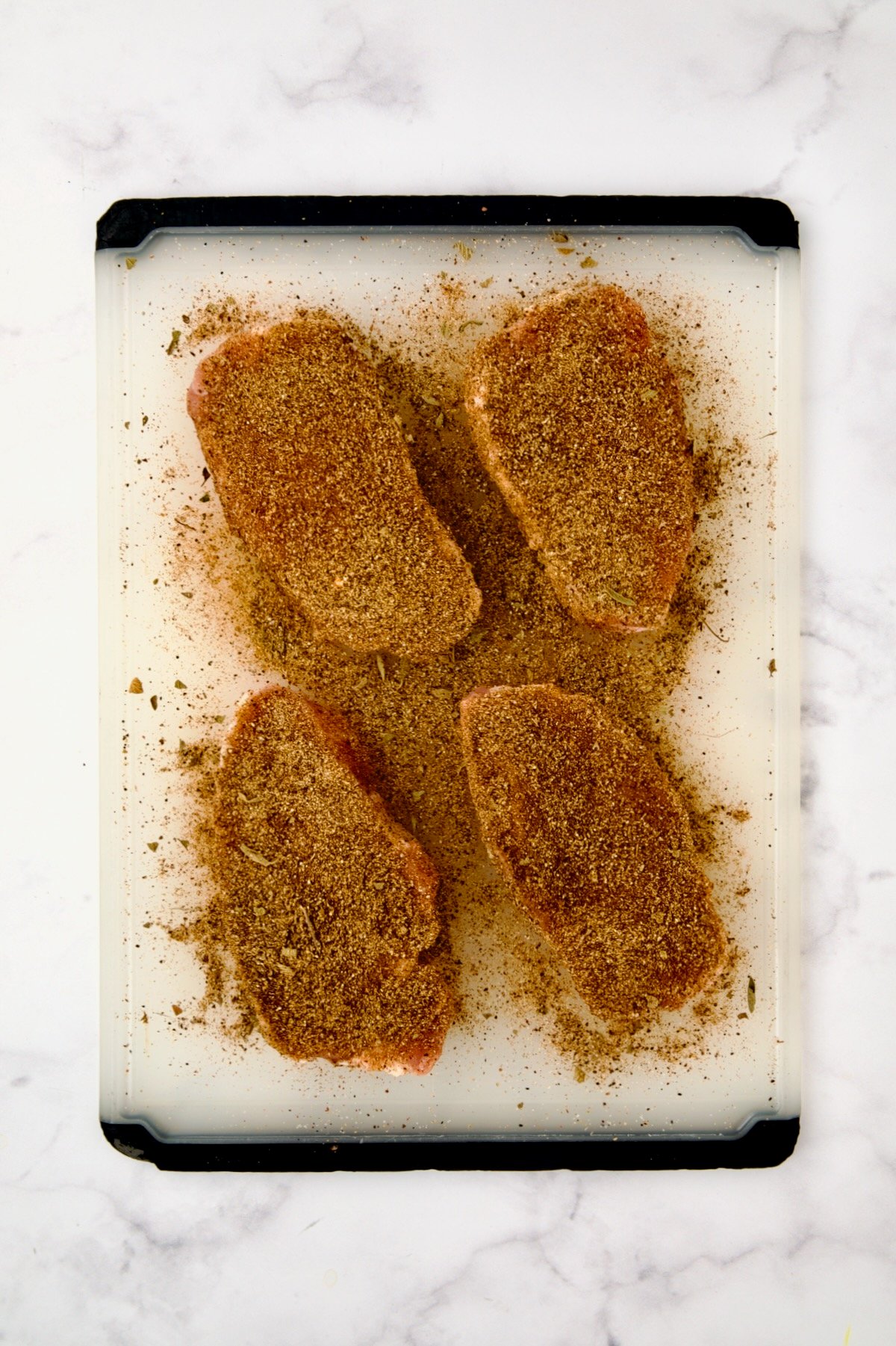 Four boneless pork chops on white plastic cutting board after being generously dusted with spice rub.