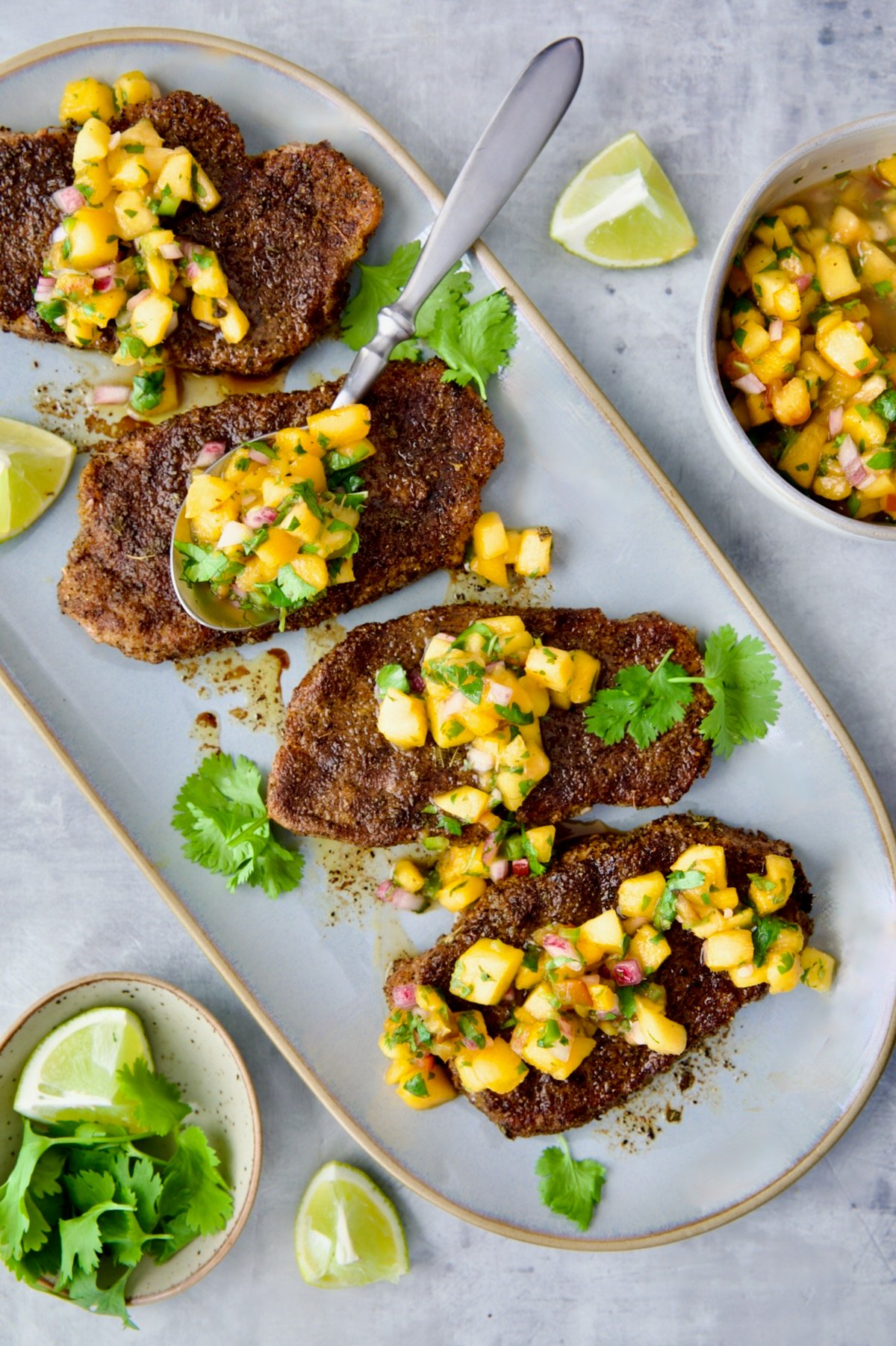Four Pork Chops with Peach Salsa on oval gray platter with spoon placing some of the salsa on each pork chop.