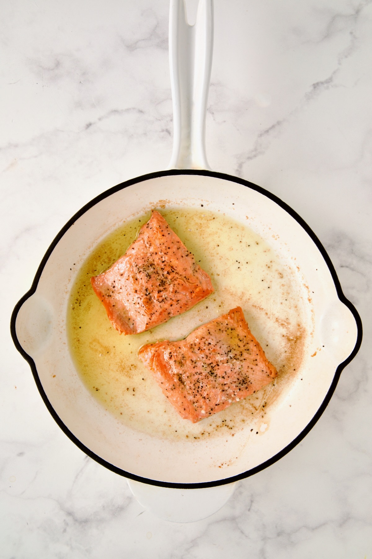 Two browned pieces of salmon in a white enamel cast iron skillet that have been browned.