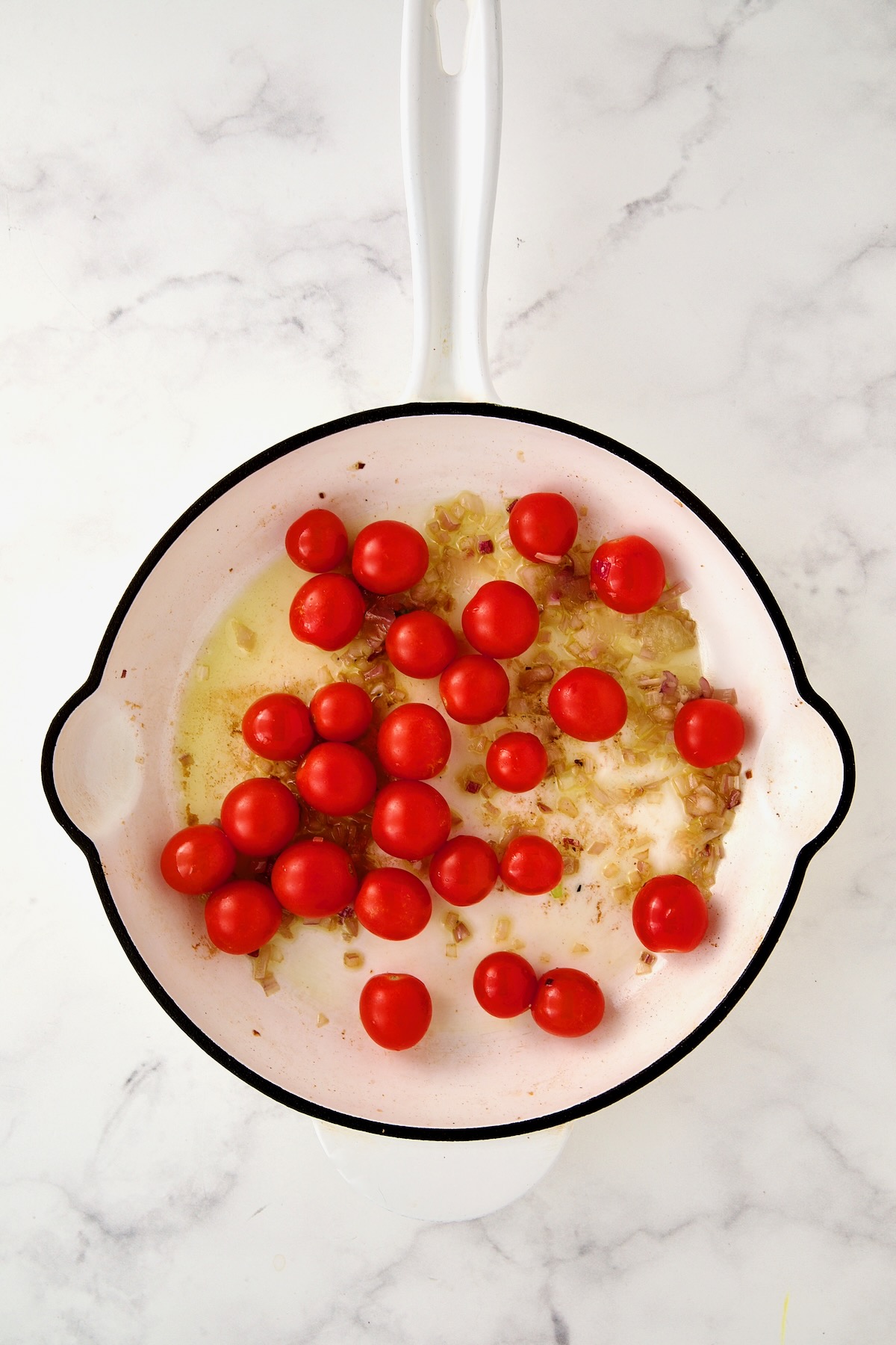 Whole cherry tomatoes added to shallots in white enamel cast iron skillet.
