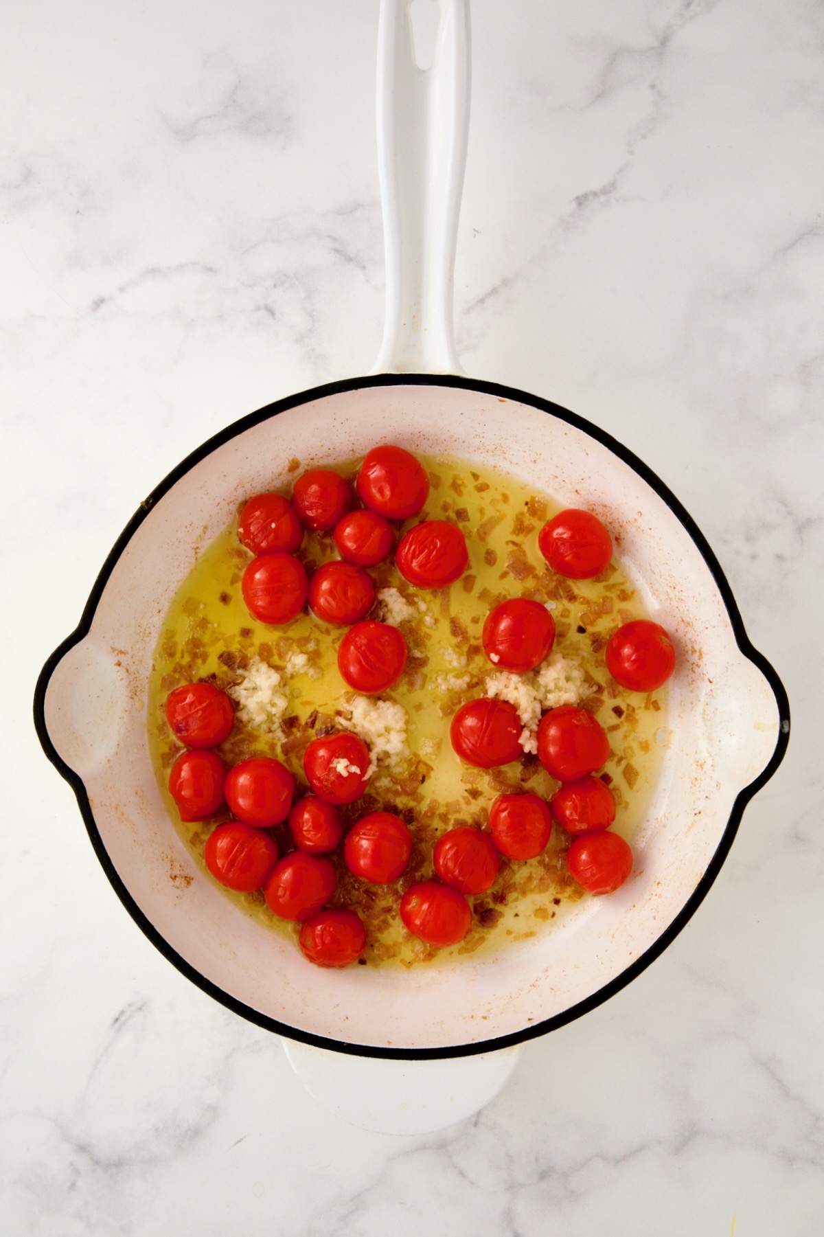 Burst cherry tomatoes and shallots in white enamel cast iron skillet with minced garlic added.