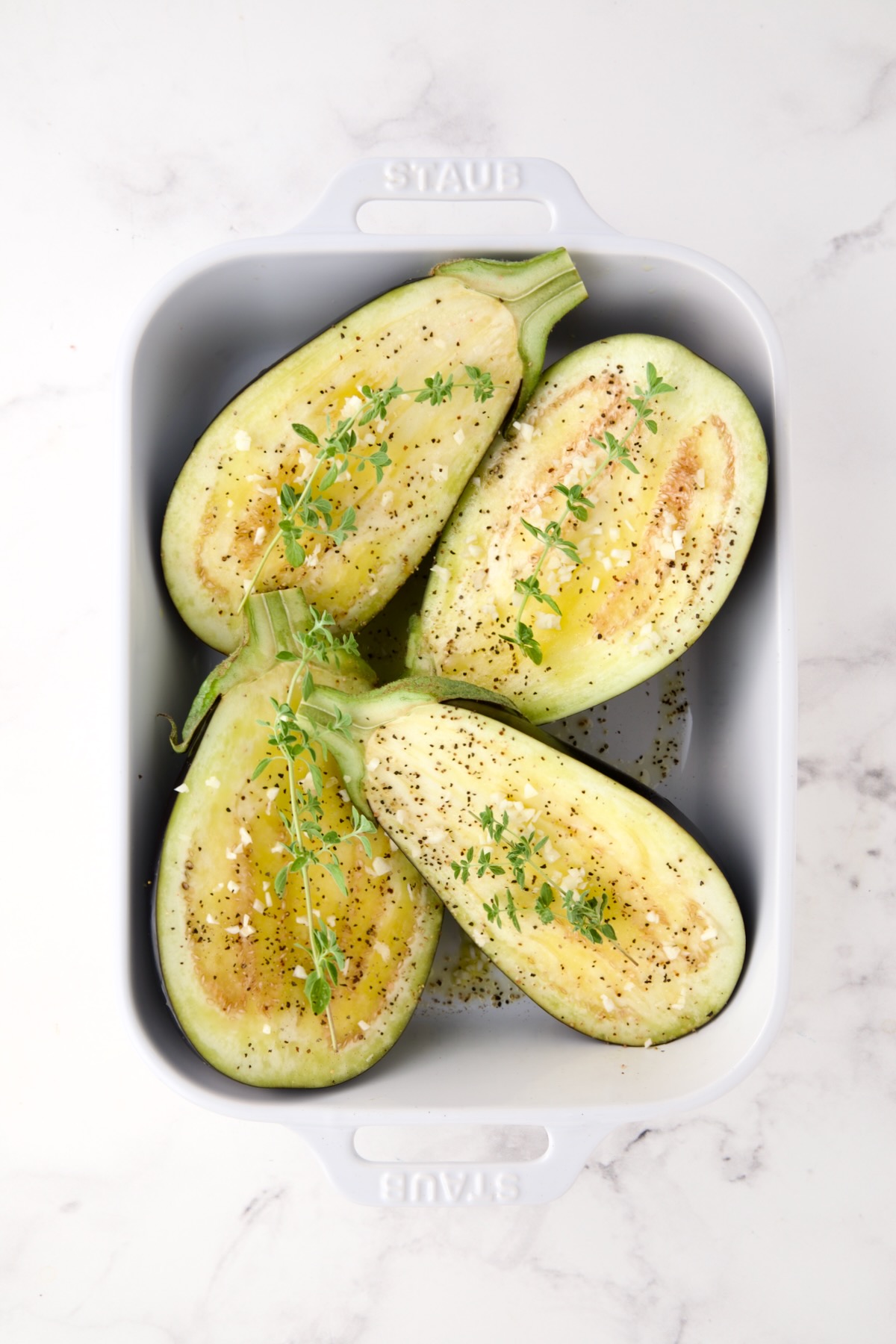 Four eggplant halves in white rectangular baking dish with fresh garlic and herbs over each half before being baked.