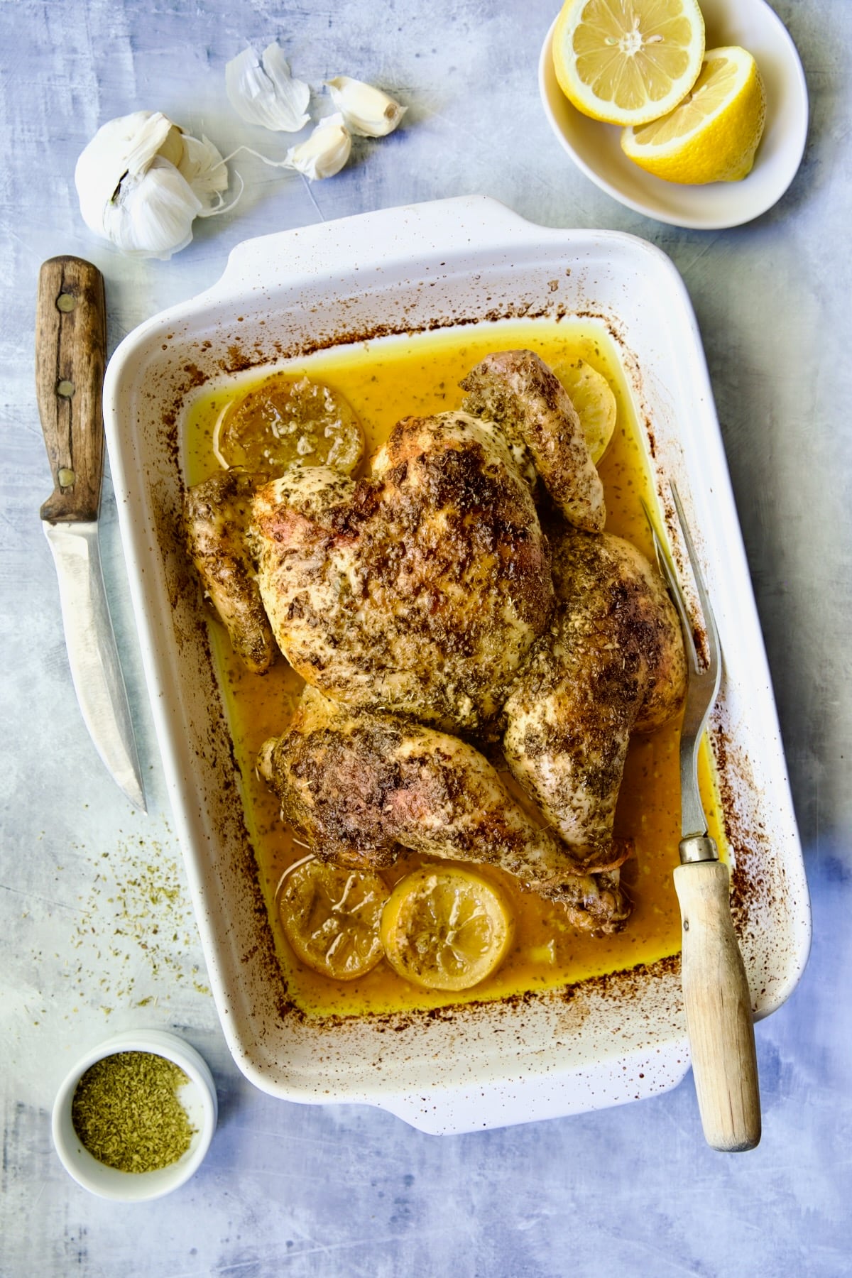 Greek Roast Chicken in white rectangular roasting pan with carving knife and meat fork.