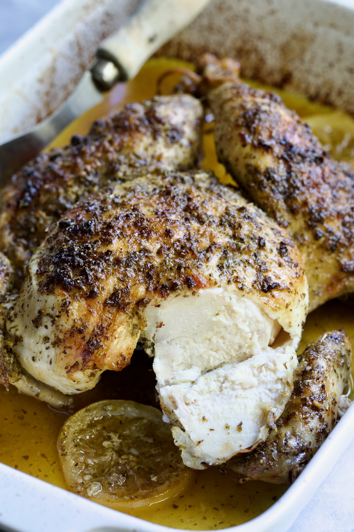 Greek Roast Chicken in white rectangular roasting pan with breast sliced into.
