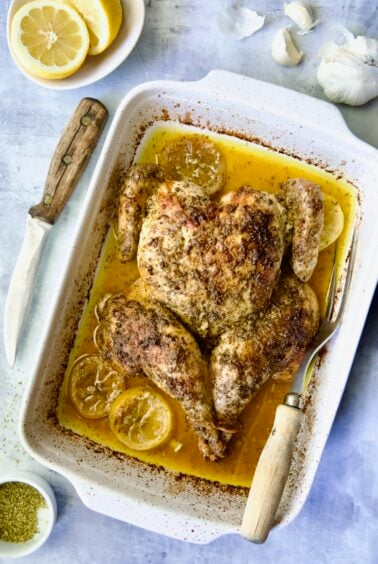 Greek Roast Chicken in white rectangular roasting pan with carving knife and meat fork.