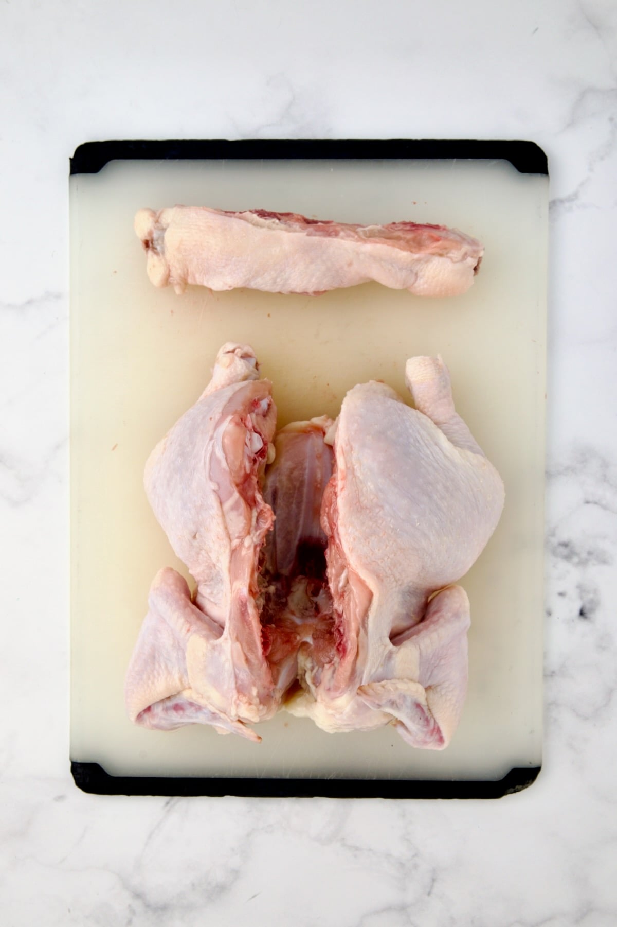 Fryer-size chicken breast side down on white plastic cutting board with backbone cut out.