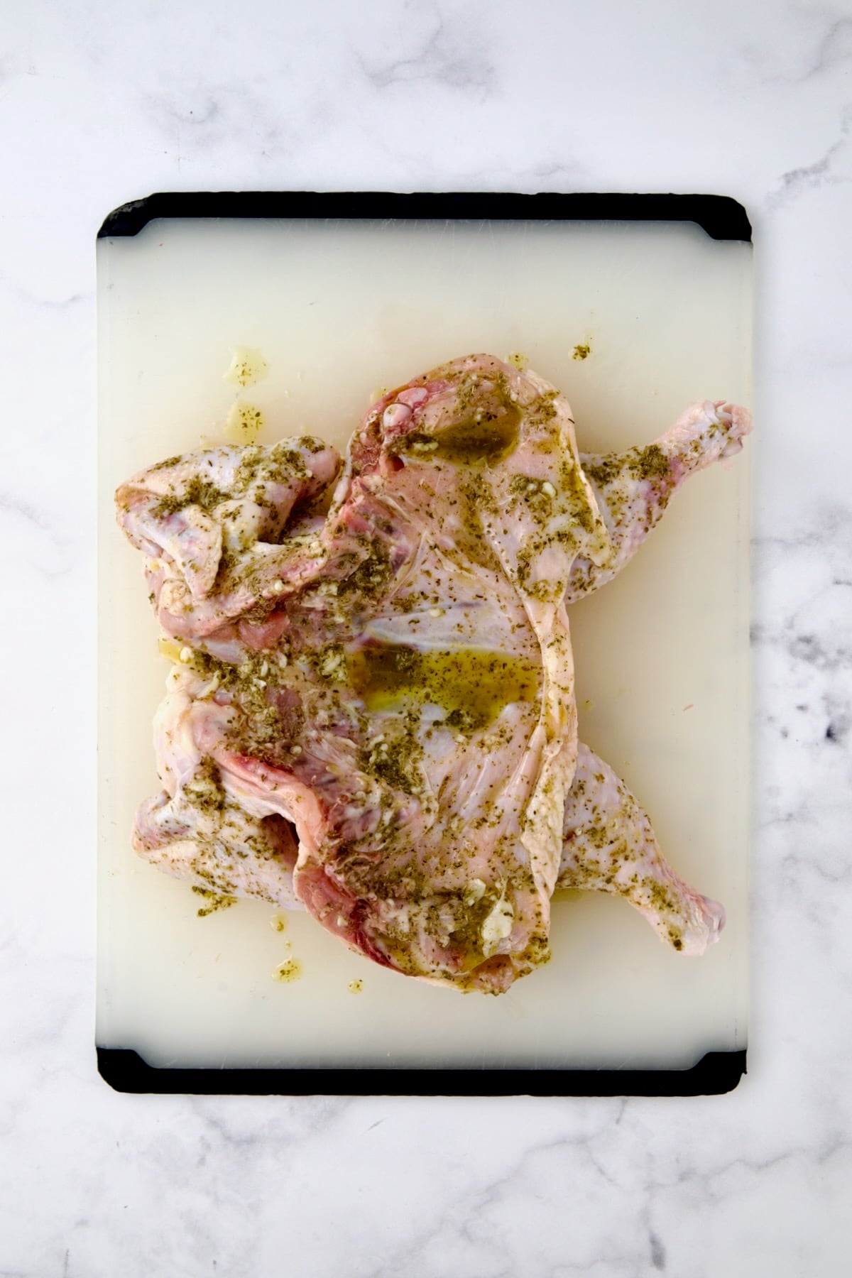Spatchcocked fryer-size chicken on white plastic cutting board smothered in olive oil, lemon juice, garlic and herbs.
