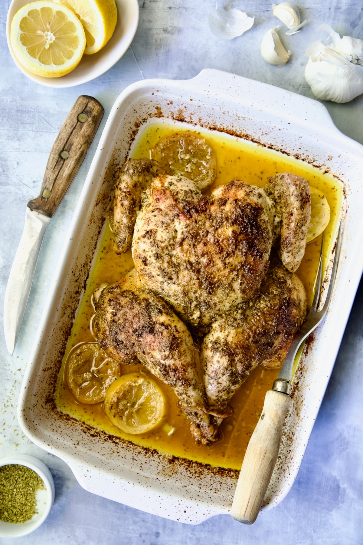 Greek Roast Chicken in white rectangular roasting pan with carving knife and meat fork.