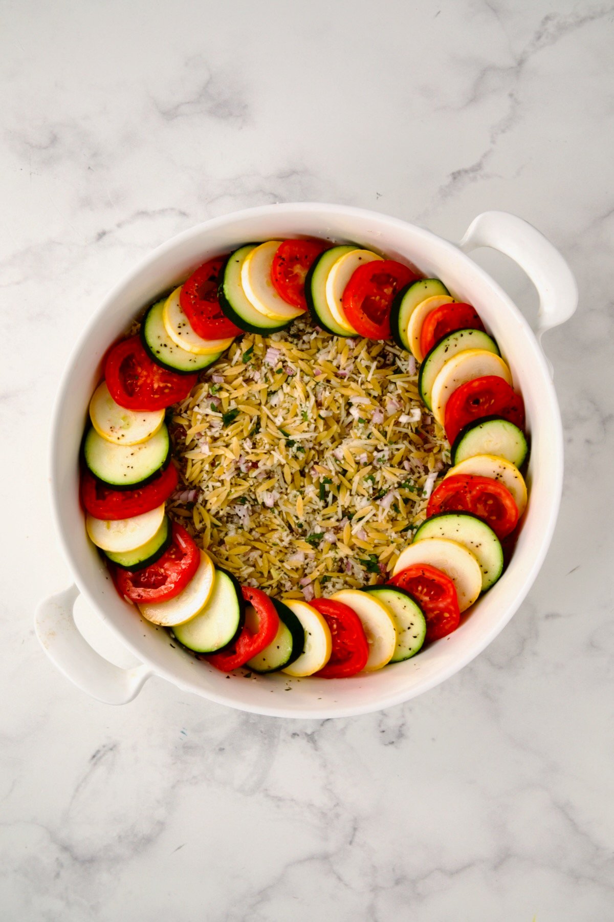 Ingredients for tian in round white baking dish with one circle of yellow squash, zucchini and tomatoes.