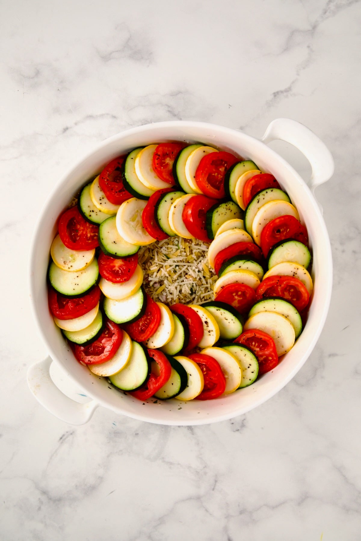 Ingredients for tian in round white baking dish with two circles of yellow squash, zucchini and tomatoes.