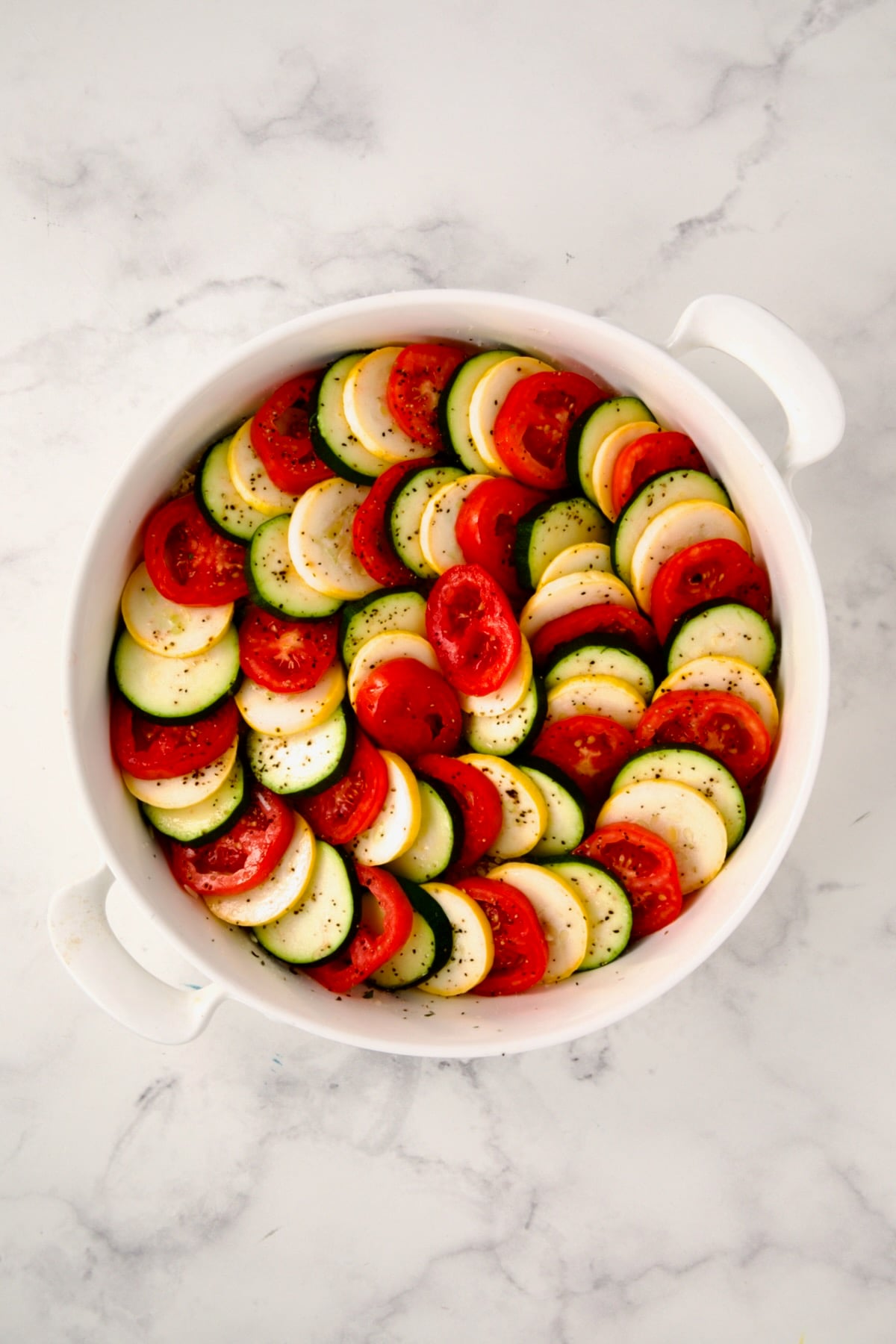 Ingredients for tian in round white baking dish with all the yellow squash, zucchini and tomatoes arranged on top.