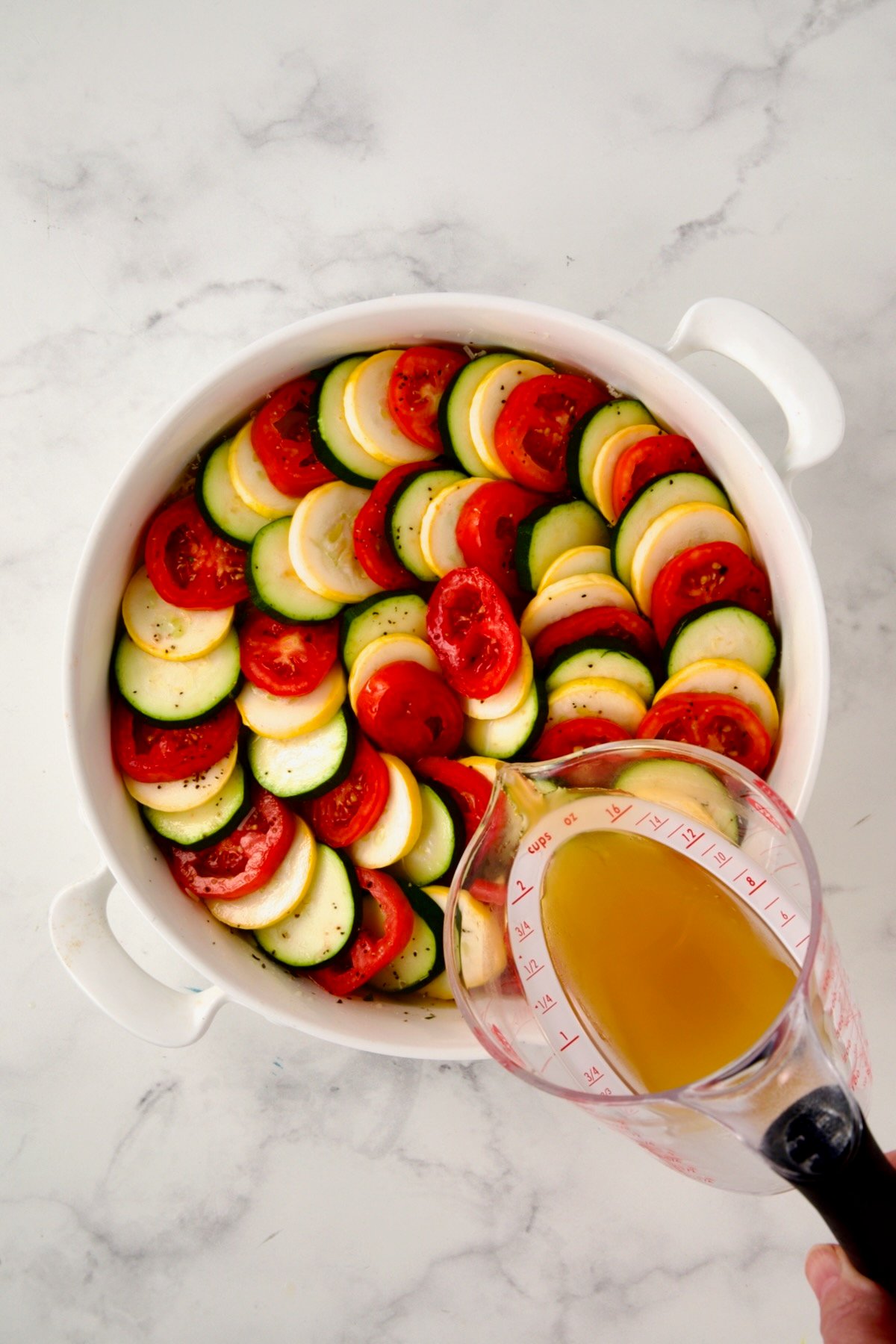 Chicken broth being poured over arranged tian in round white baking dish.