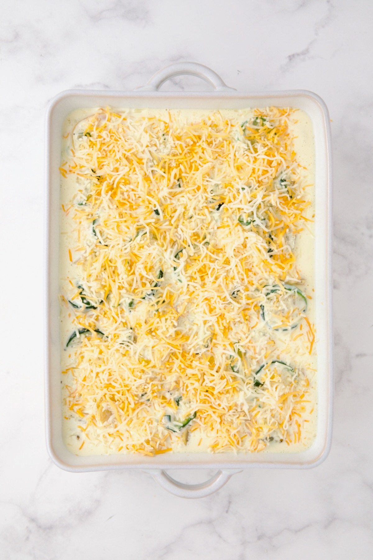Cooked zucchini, yellow squash, poblano, and corn in white rectangular baking dish with custard added and cheese sprinkled over the top.