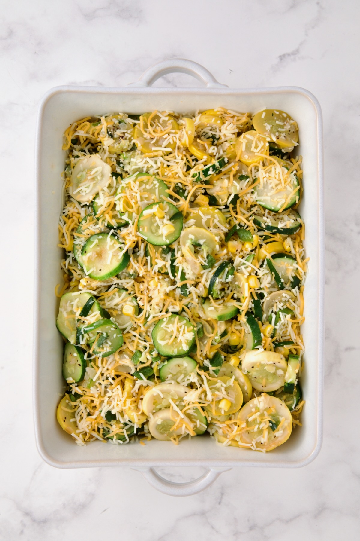 Cooked zucchini, yellow squash, poblano, and corn in white rectangular baking dish with shredded cheese added.