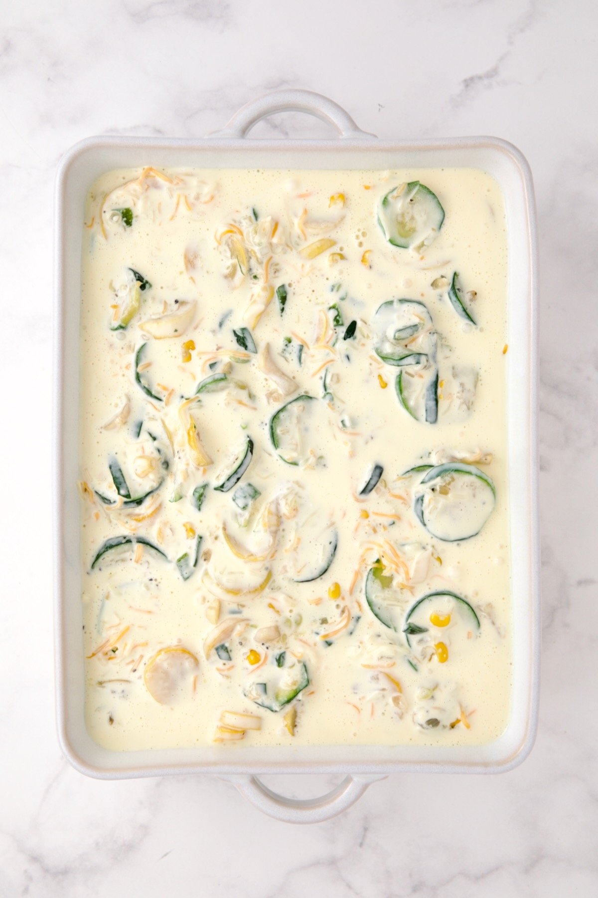 Cooked zucchini, yellow squash, poblano, and corn in white rectangular baking dish with shredded cheese and custard added.