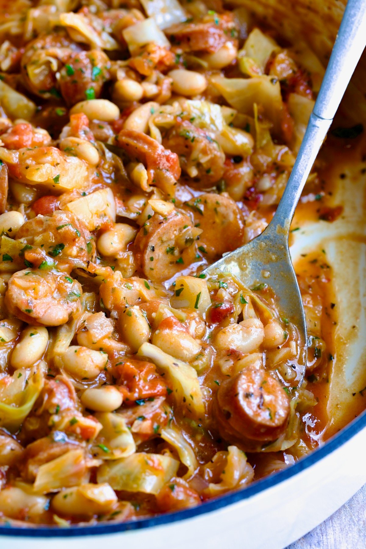 Cabbage Sausage White Bean Skillet in a white enamel cast iron pan with a serving spoon scooping some up.