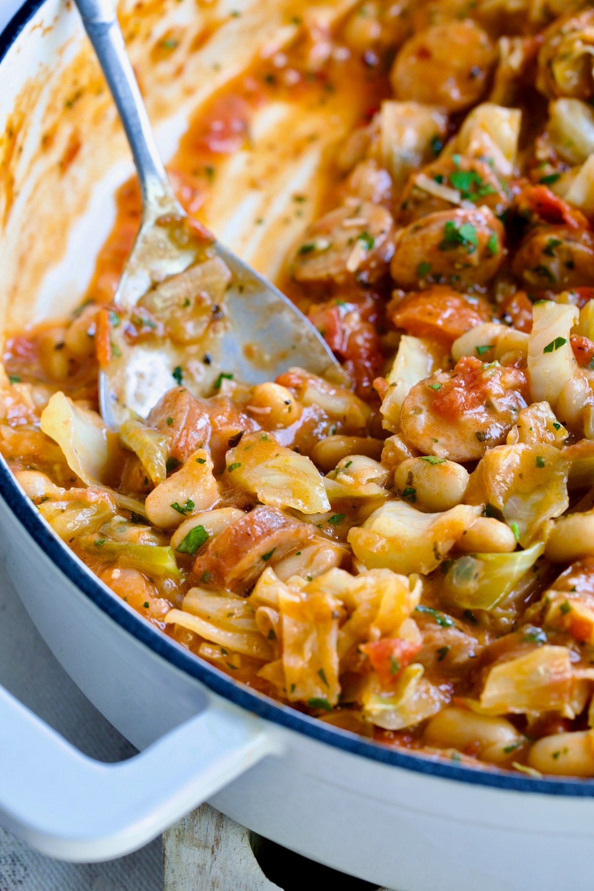 Cabbage Sausage White Bean Skillet in a white enamel cast iron pan with a serving spoon scooping some up.