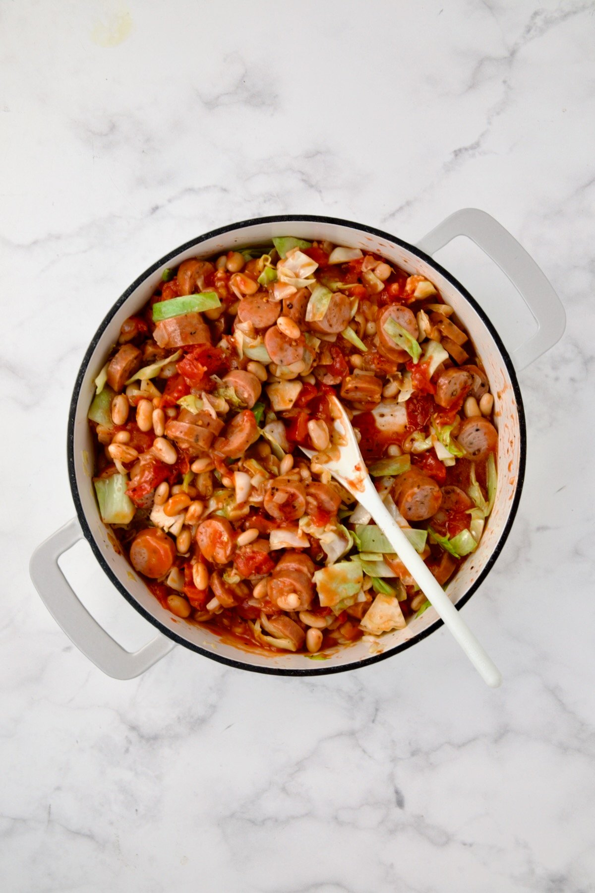 Ingredients for Cabbage Sausage White Bean Skillet being sauteed with white spoon in white enamel cast iron skillet.