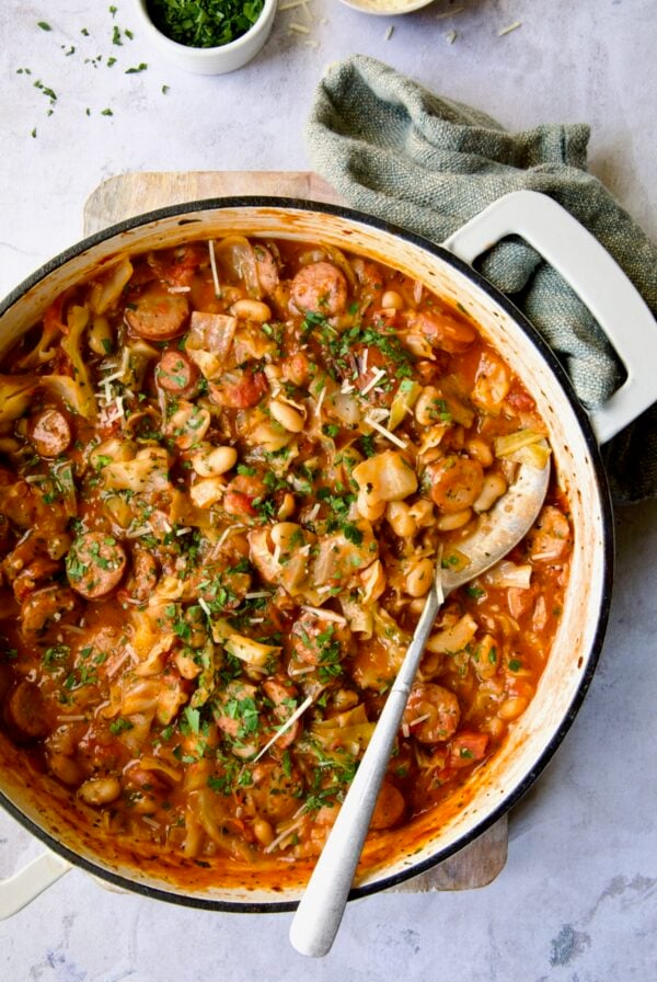 Cabbage Sausage White Bean Skillet in a white enamel cast iron pan with a serving spoon.