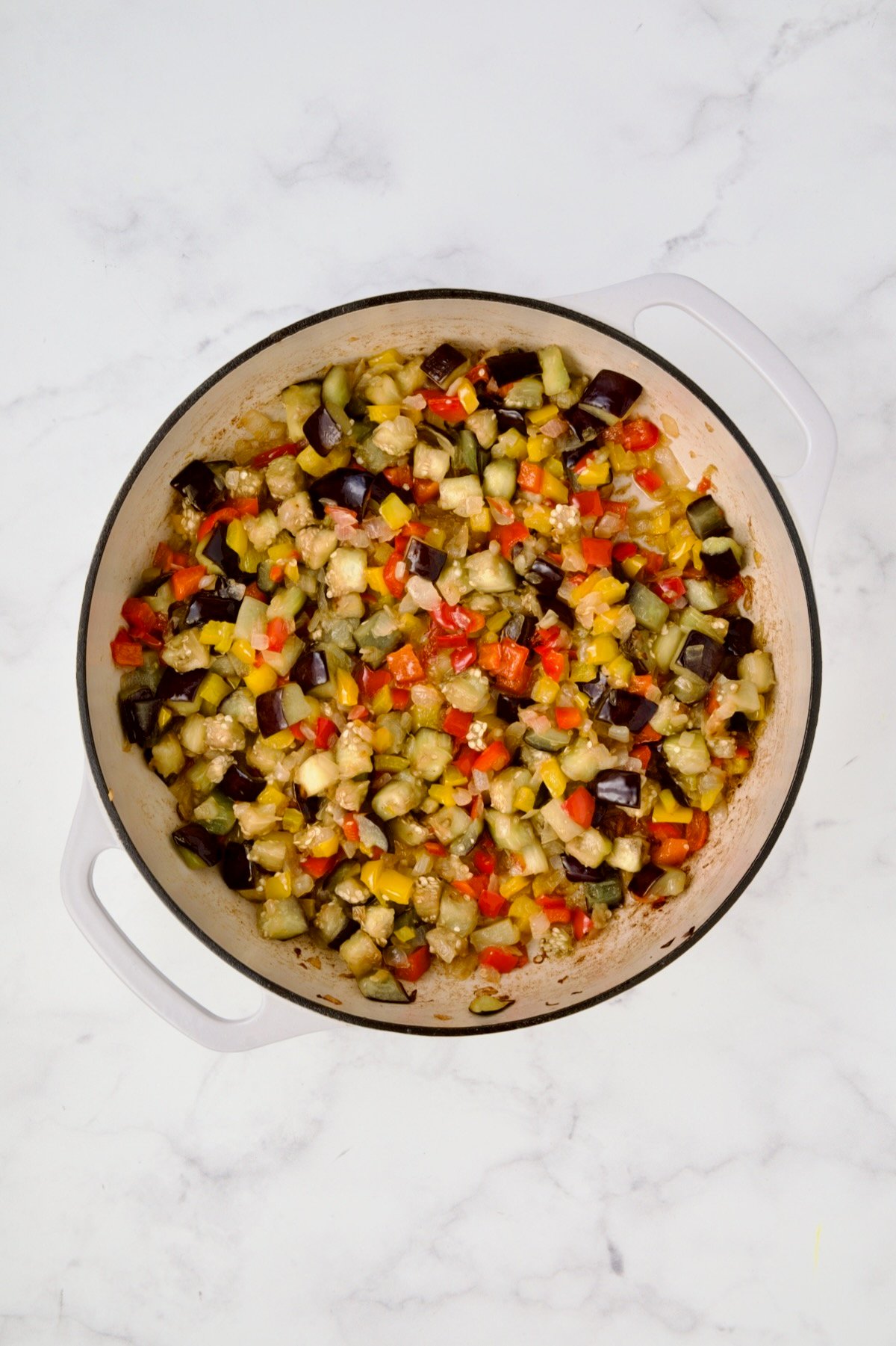 Cooked yellow bell peppers, red bell peppers, eggplant, and onions in white enamel cast iron skillet.