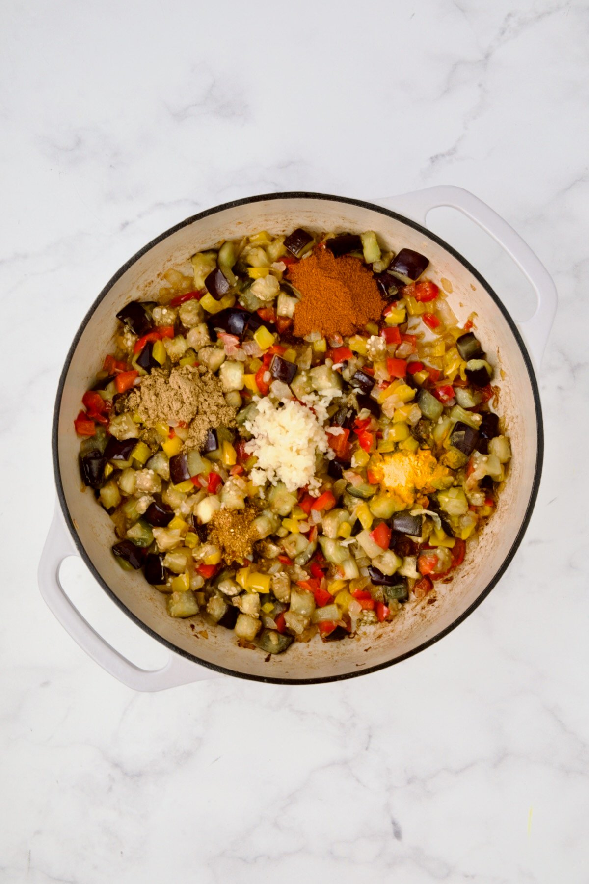 Cooked yellow bell peppers, red bell peppers, eggplant, and onions in white enamel cast iron skillet with garlic and spices added.